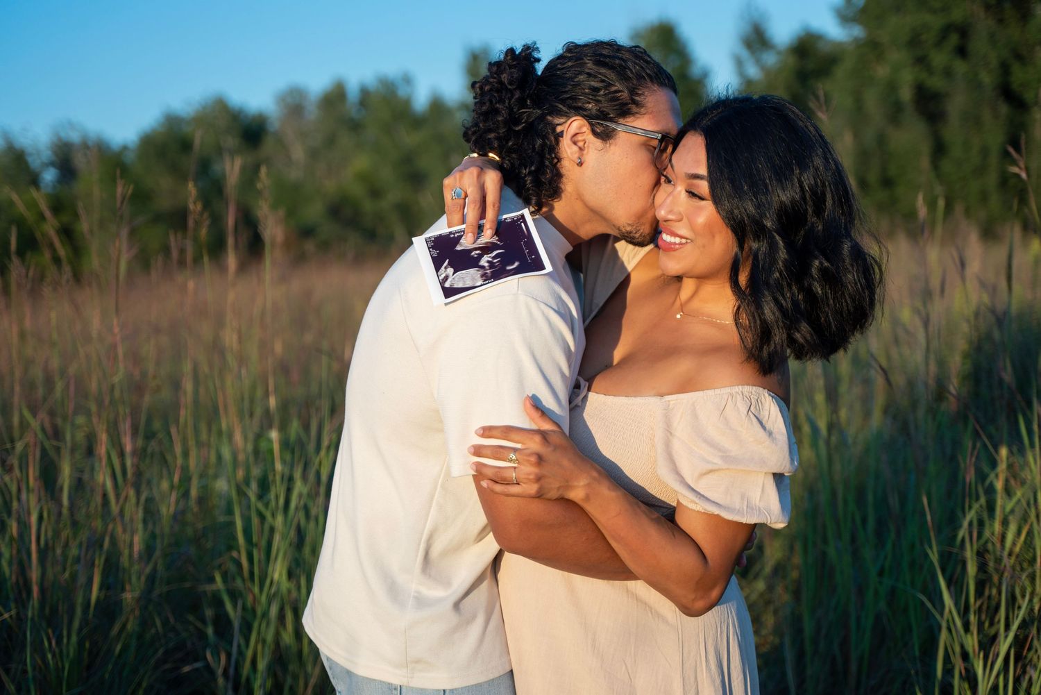 Dad to be kisses mom to be in glowy sunshine in a dreamy field with blue skies.