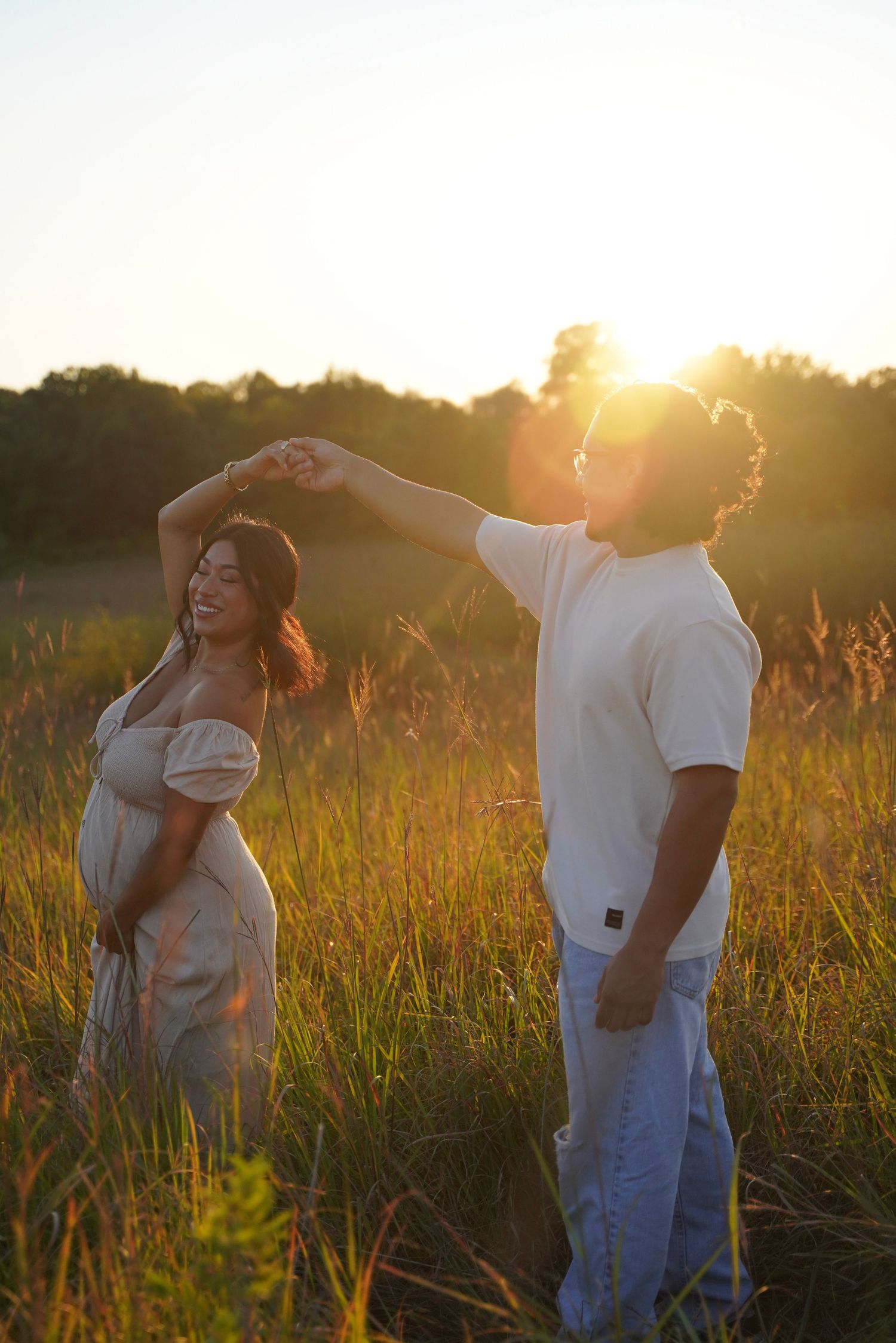 Hispanic couple maternity session in a field with sunlight at golden hour. Dad to be is twirling mom to be.