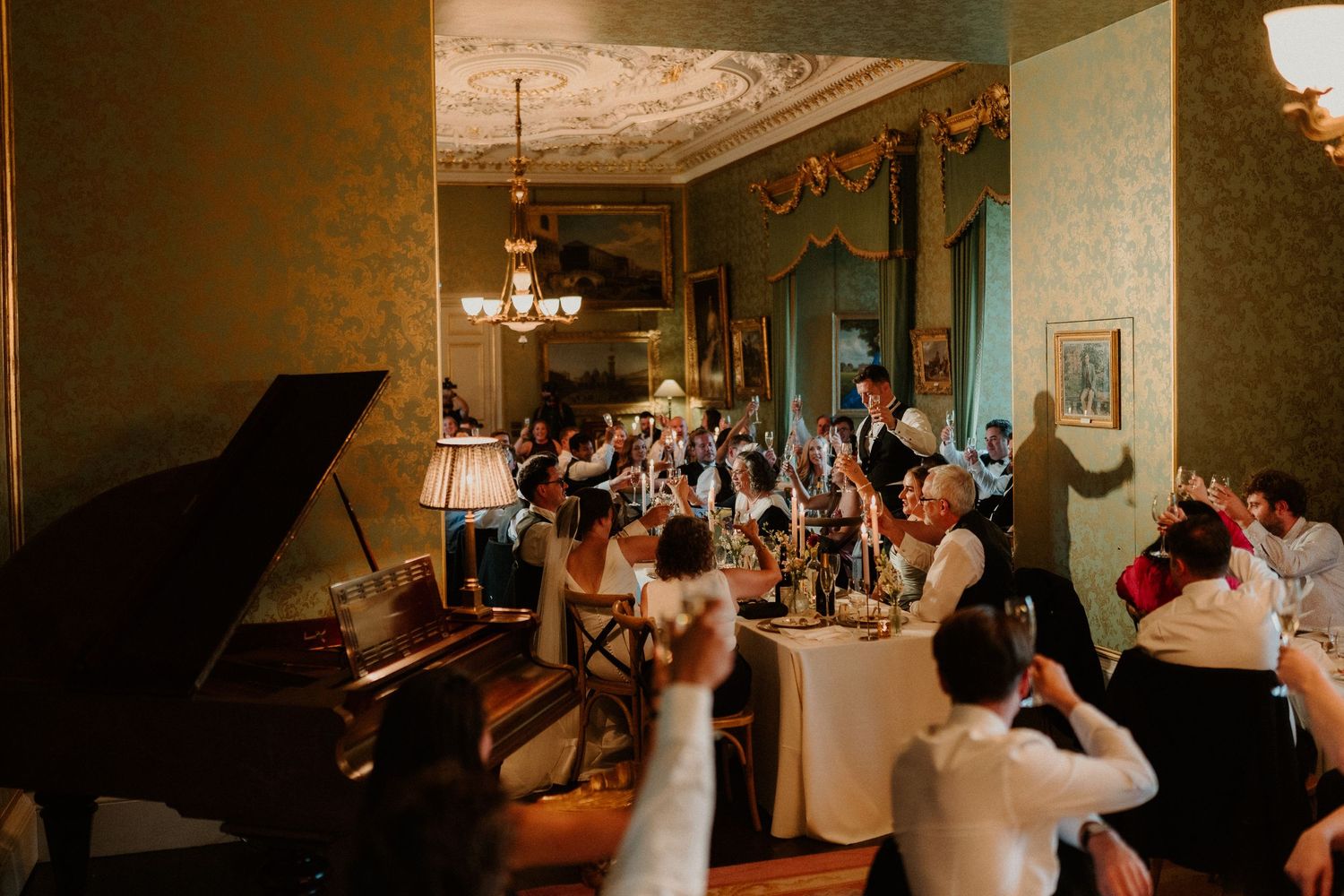 Elegant wedding reception in a grand room with chandelier, piano, and guests raising glasses for a toast.