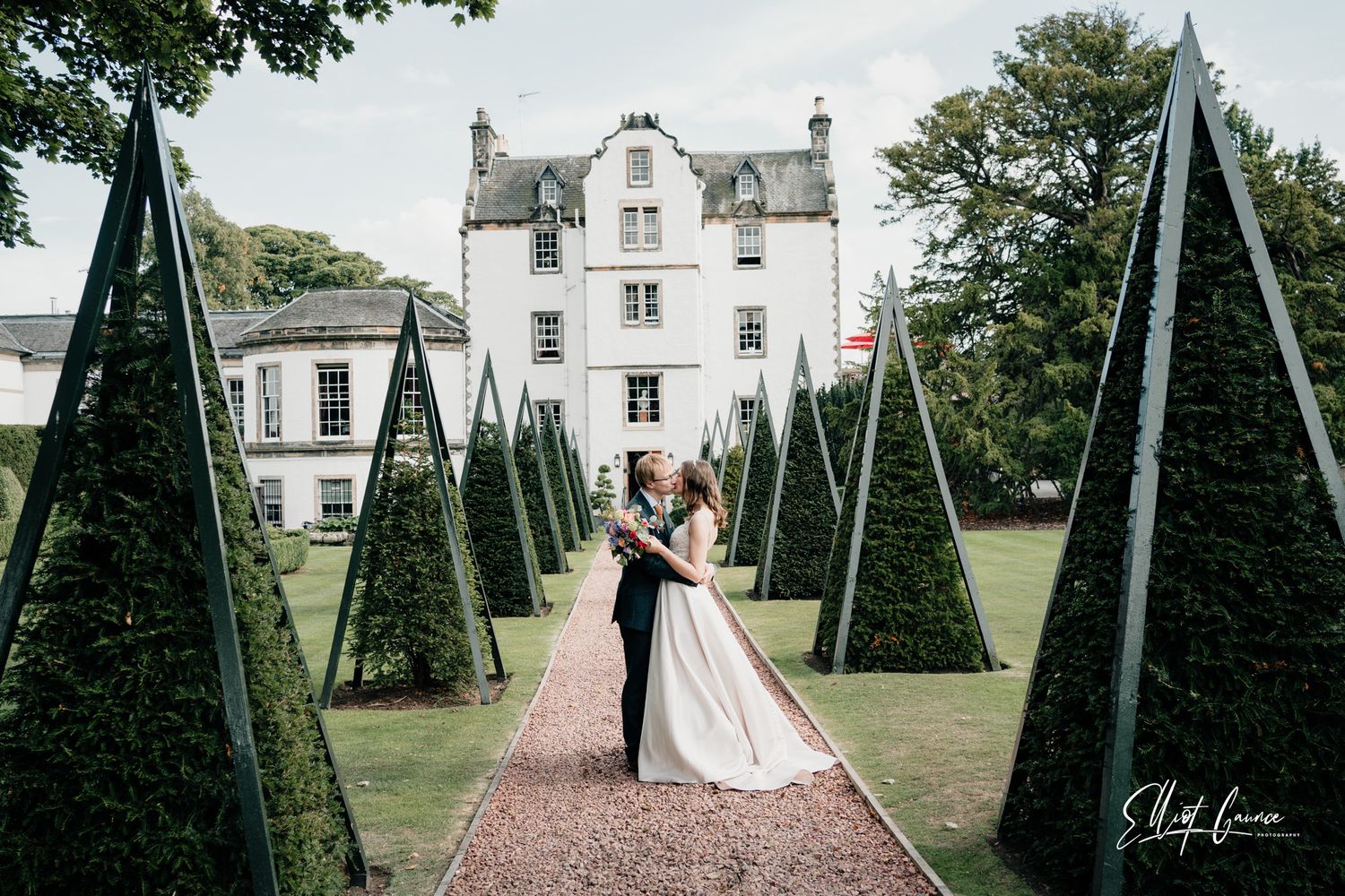 Wedding couple walks down a tree-lined garden path at an elegant white estate venue.