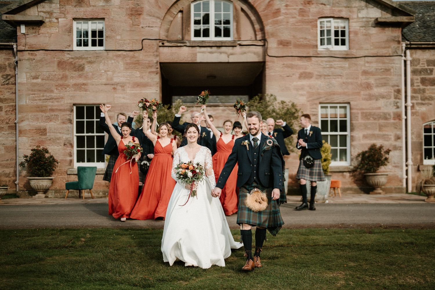 Wedding group poses in front of rustic stone building with red brick facade.