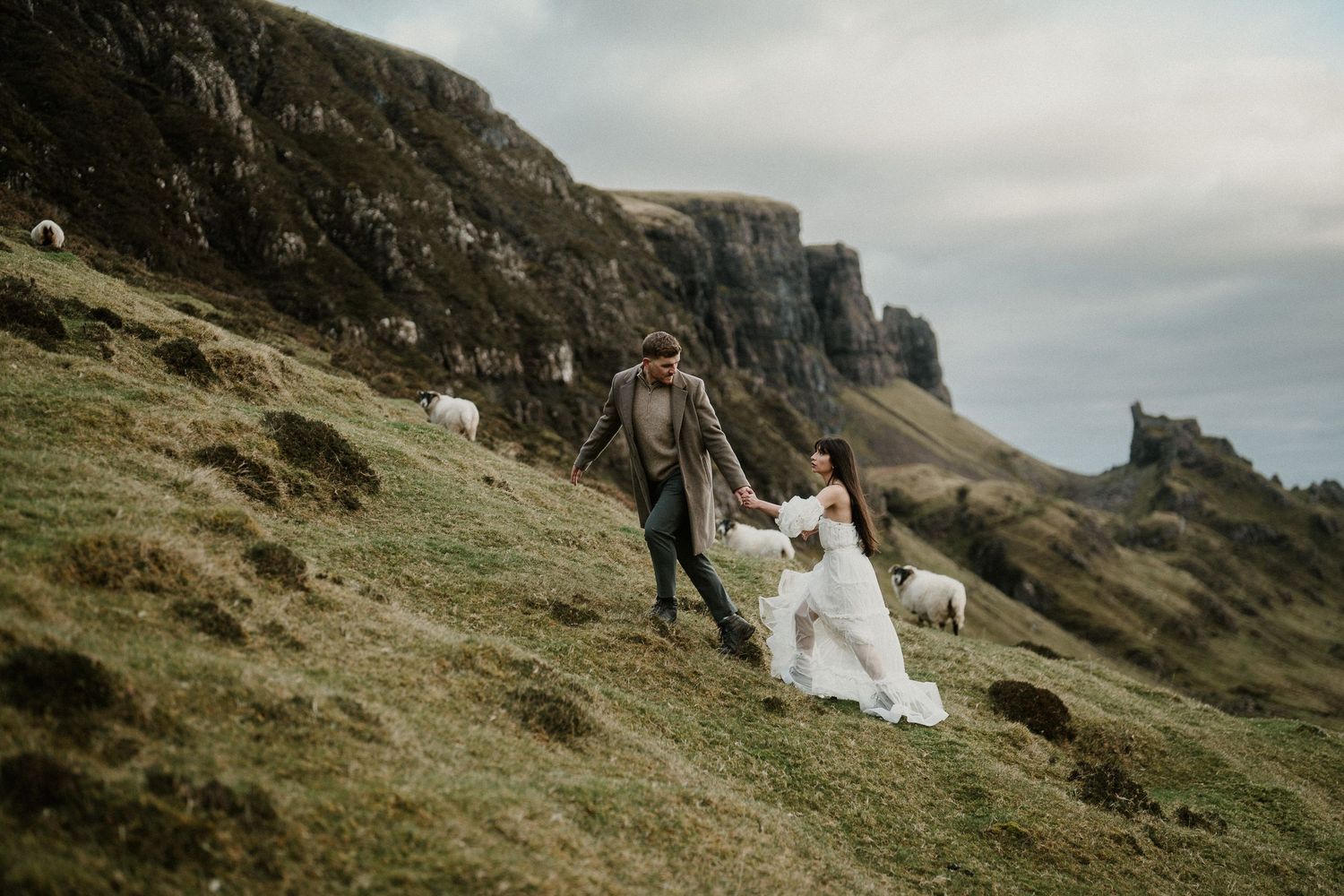 Wedding couple walks along rugged mountain path with dramatic cliffs and coastline in background.