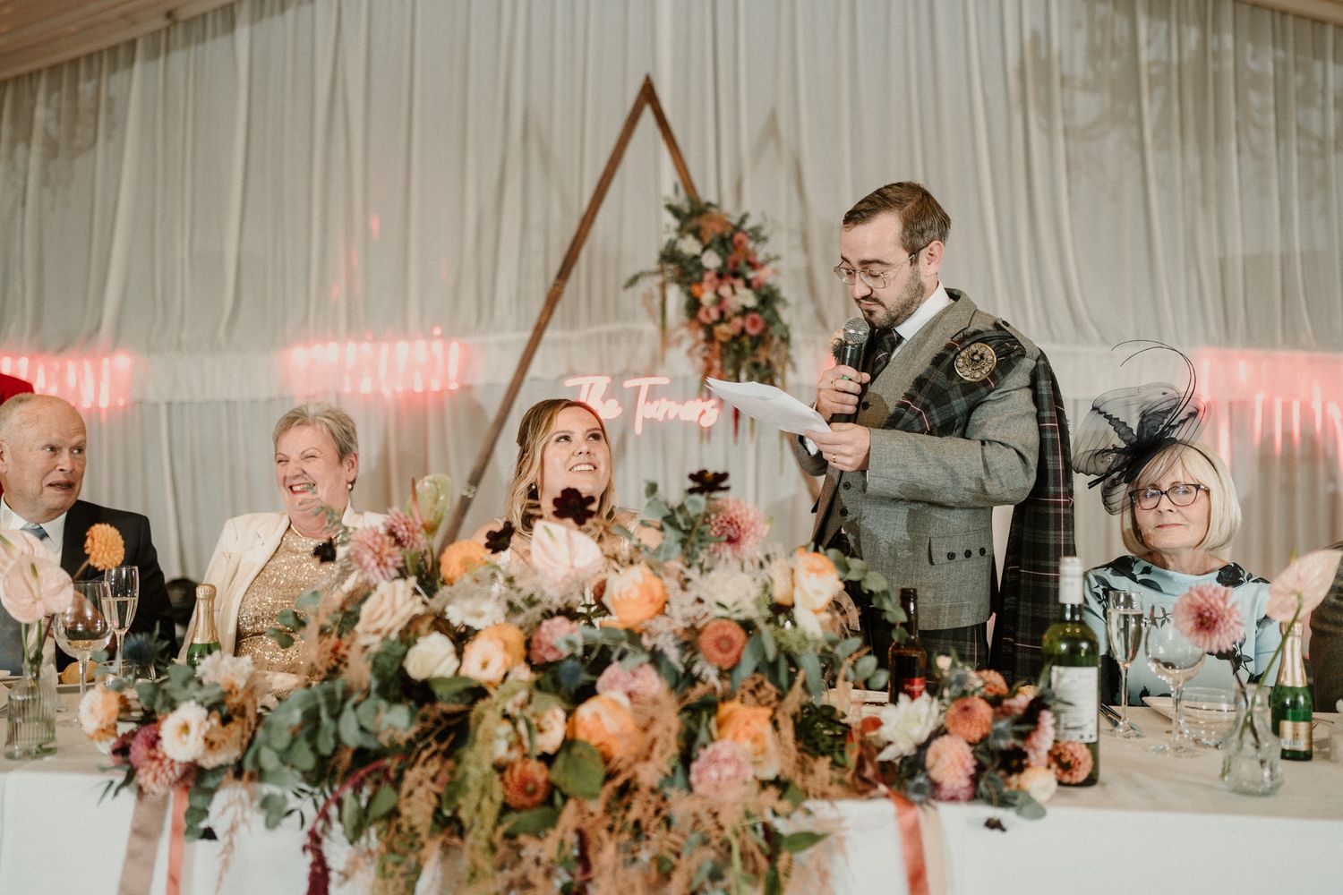 Wedding speeches at a reception with floral centerpieces and warm lighting under a tent.