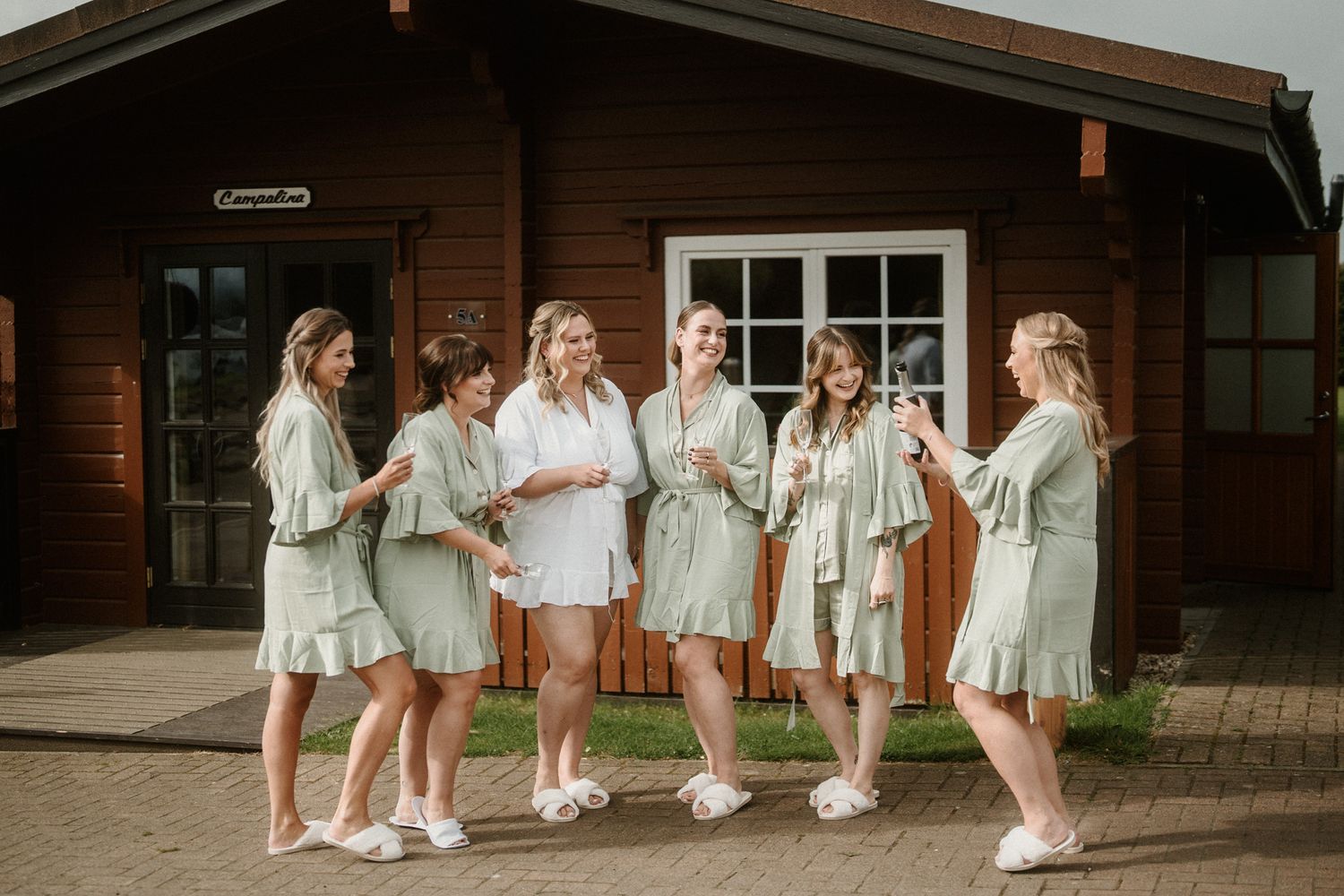 Wedding party in white dresses and sneakers poses together outside rustic wooden building.