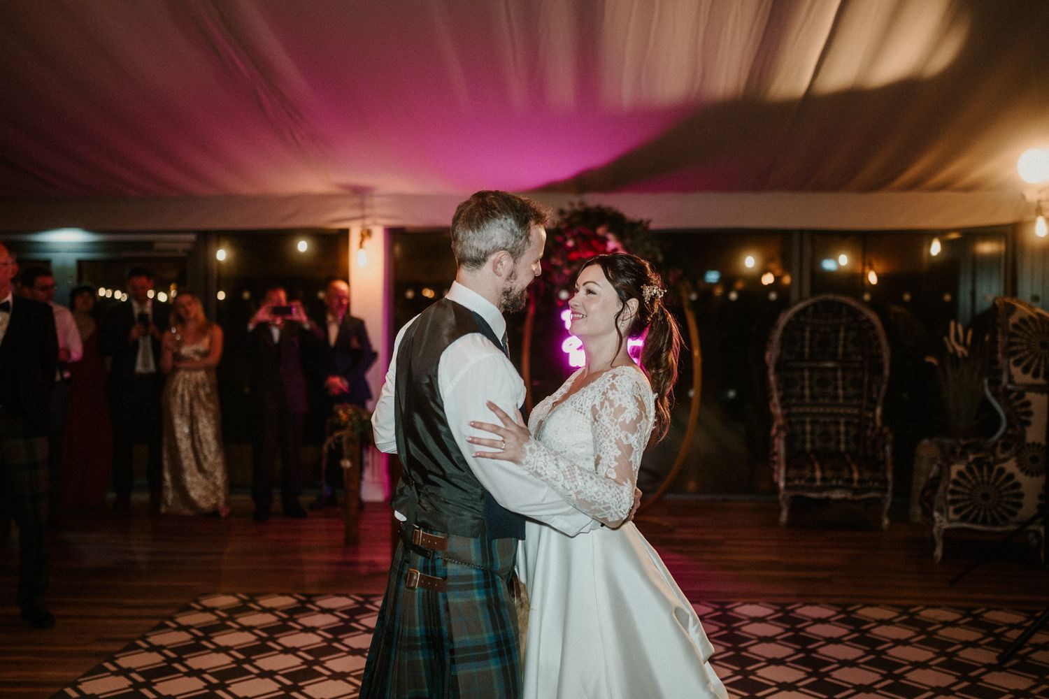 A couple shares their first dance at a Scottish wedding reception under pink mood lighting.