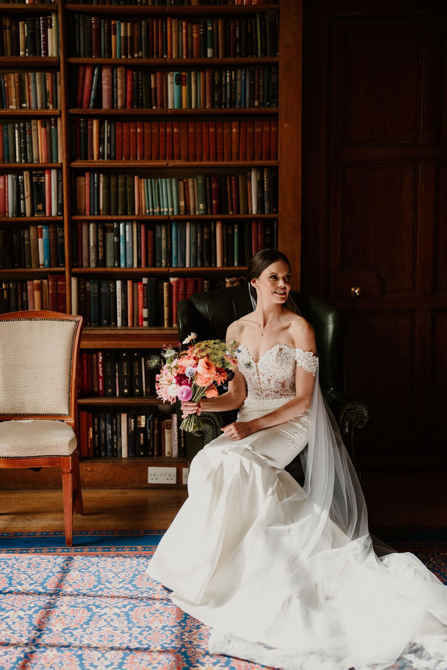 Bride in lace dress sits with floral bouquet surrounded by library bookshelves.