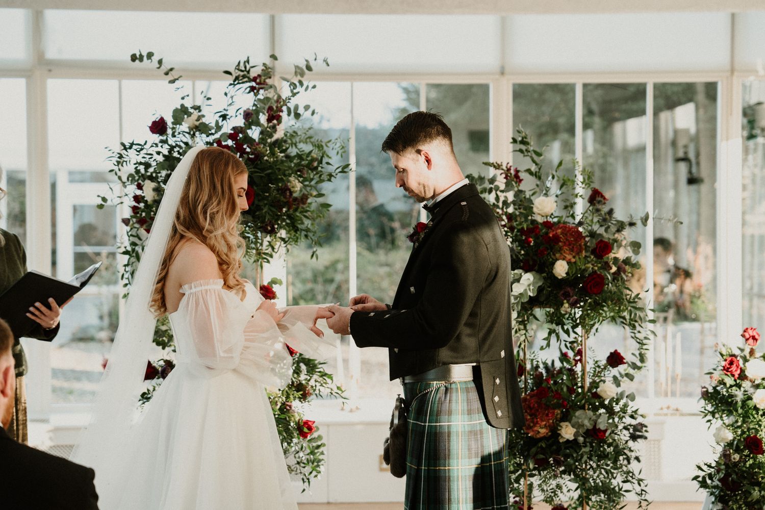 A couple exchanges rings during their wedding ceremony surrounded by floral arrangements.