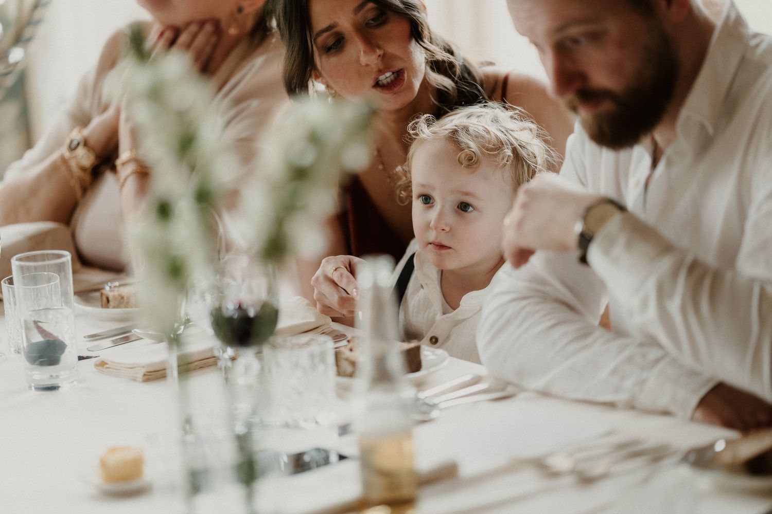 A toddler sits at an elegant wedding reception table with adults, surrounded by floral centerpieces and glassware.