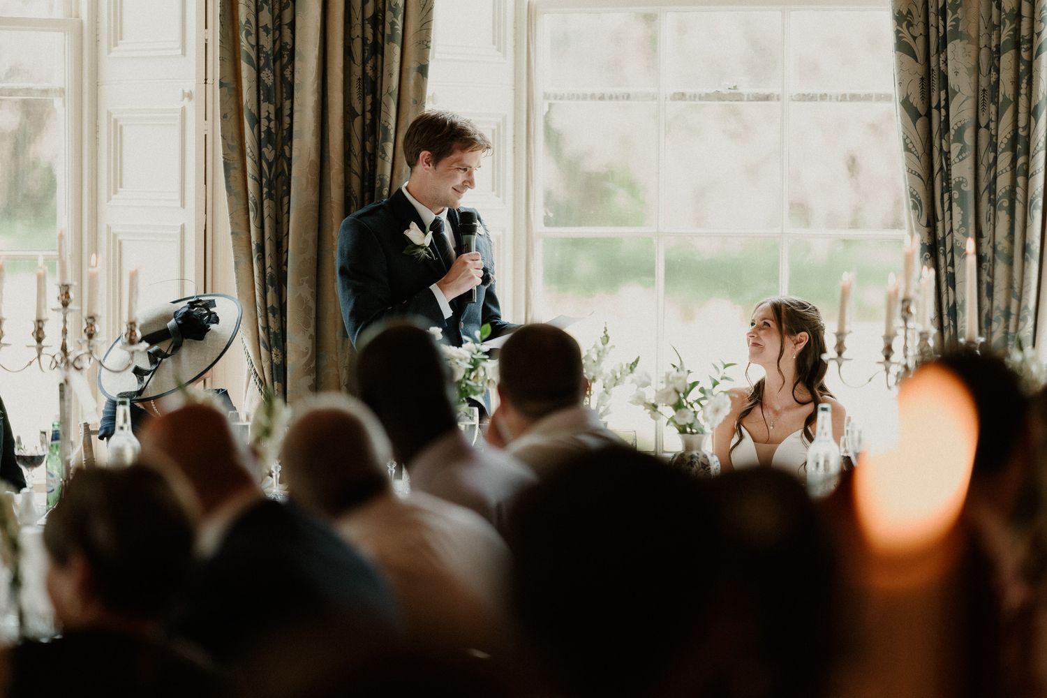 Groom giving a heartfelt wedding speech at elegant reception while bride smiles adoringly at candlelit table.