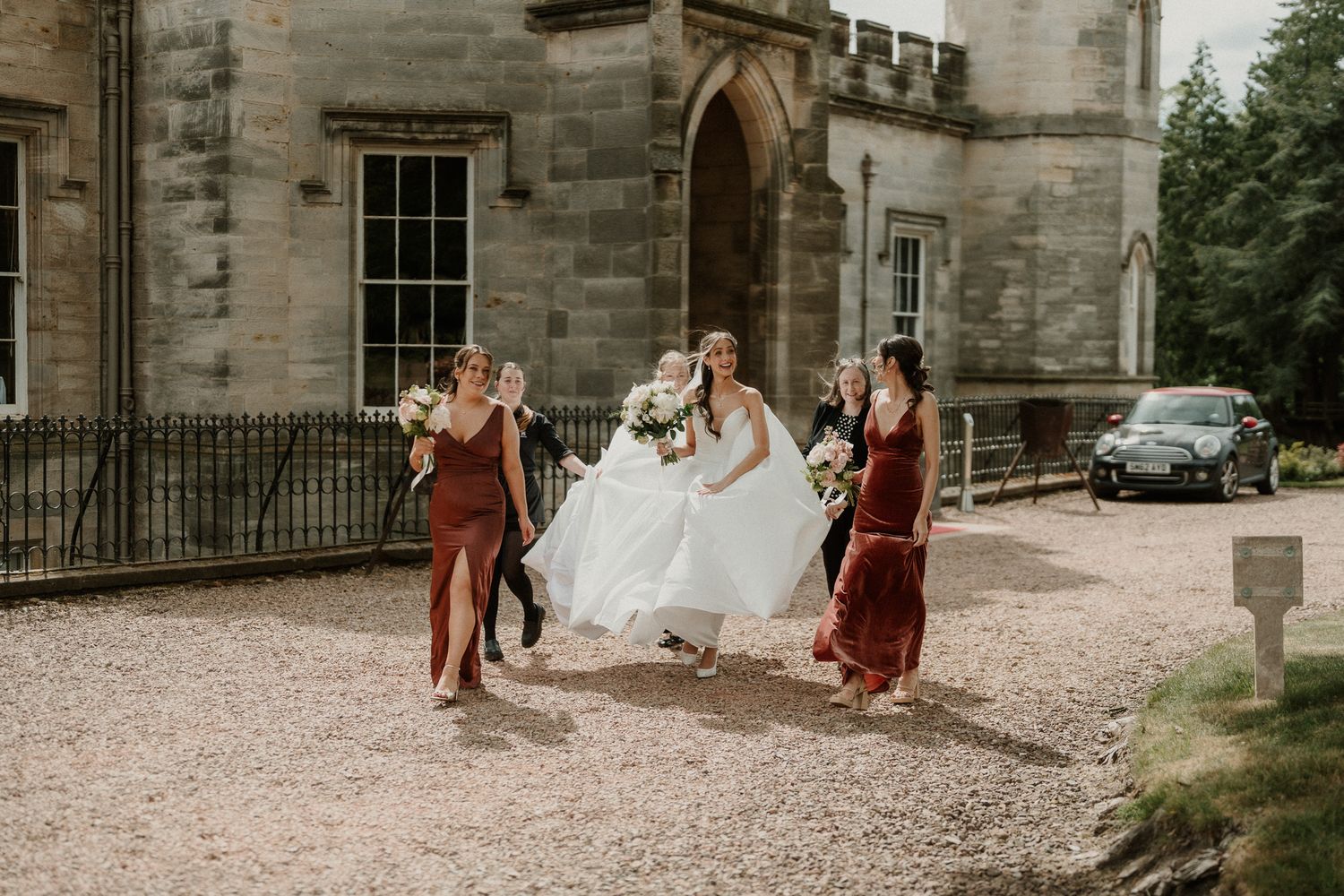 Bride in white gown walks with bridesmaids in burgundy dresses outside a stone castle venue.