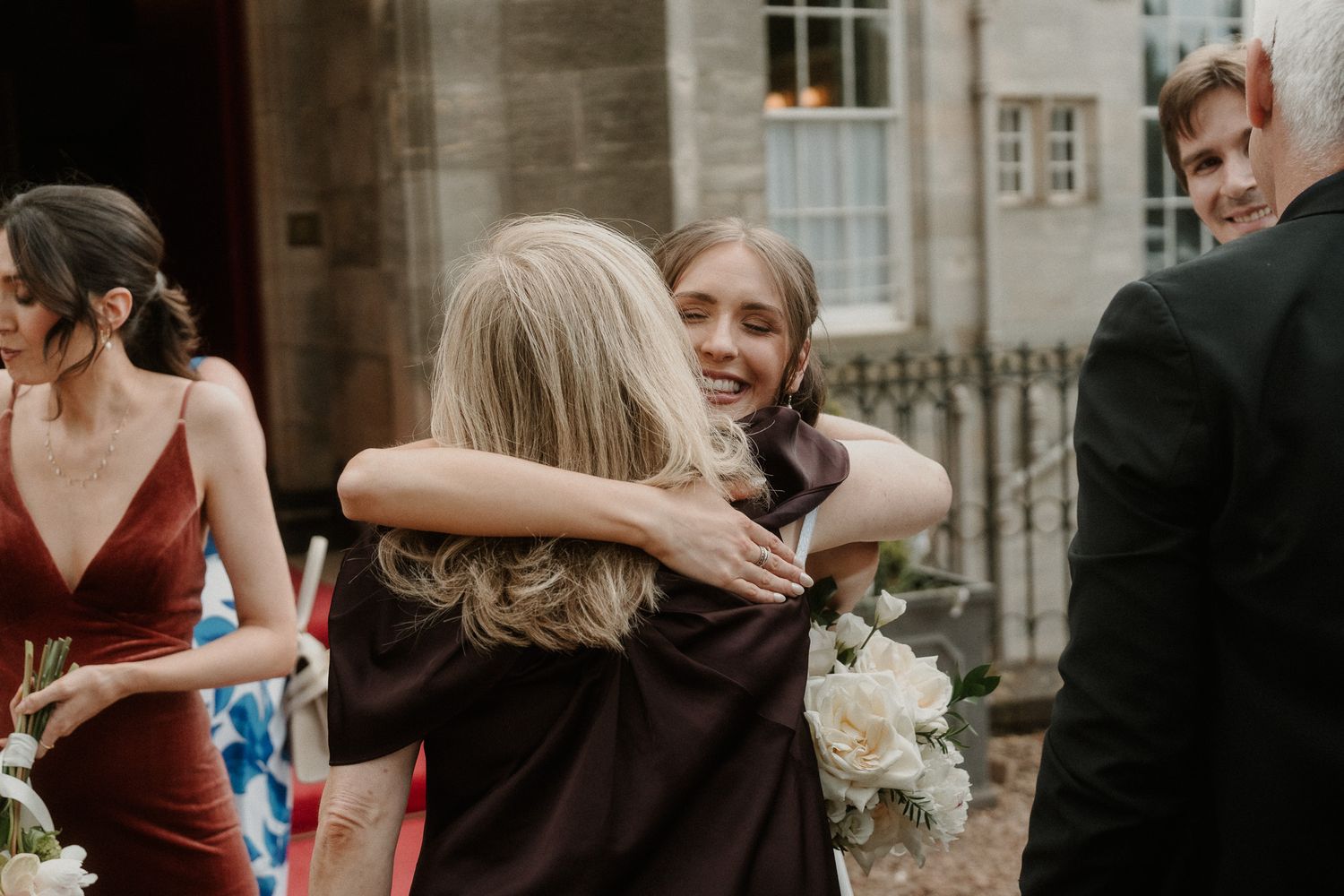 Bride hugging a guest at a wedding reception outside a stone building, with bridesmaids in burgundy dresses nearby.
