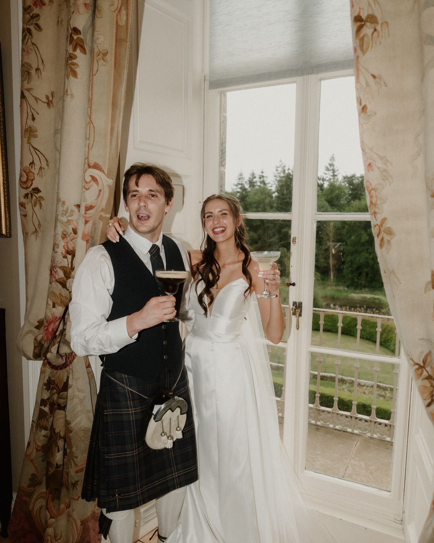 A groom in a kilt and bride in a white gown smile together holding drinks by an open French door.
