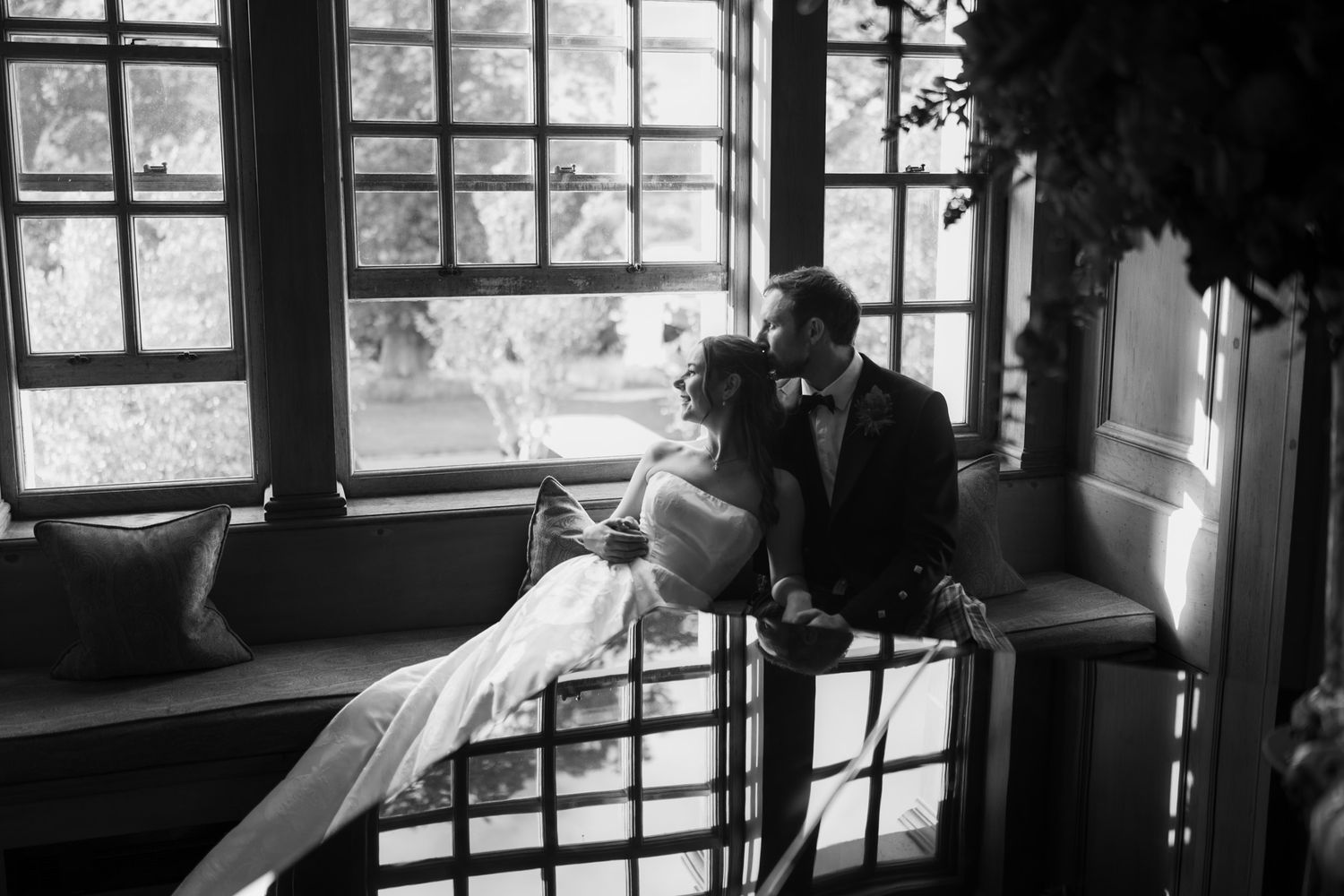 Black and white wedding photo of bride and groom sharing intimate moment by ornate windows in elegant venue.