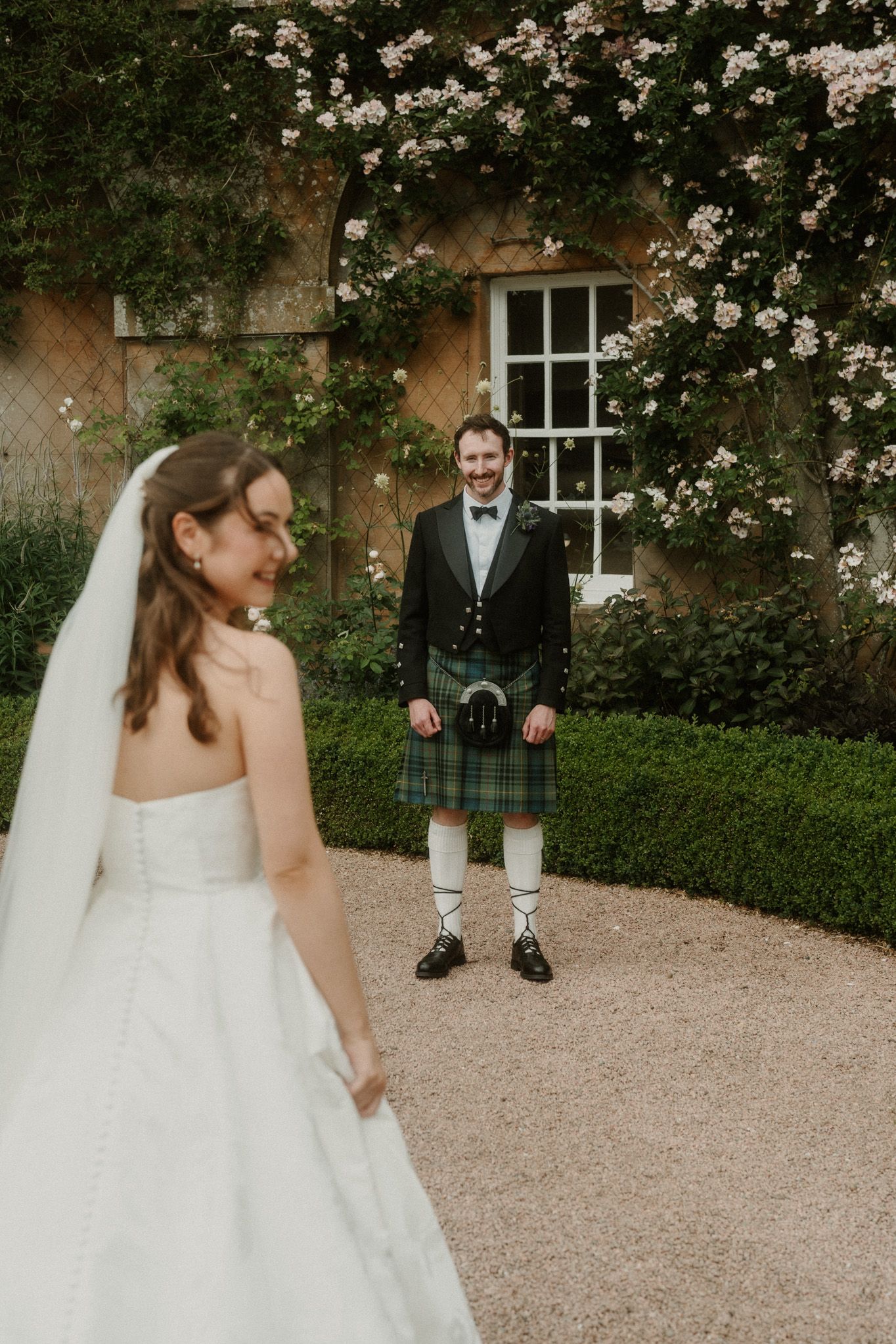 A bride in a white gown and groom in a green tartan kilt share a first look moment in a romantic garden setting.