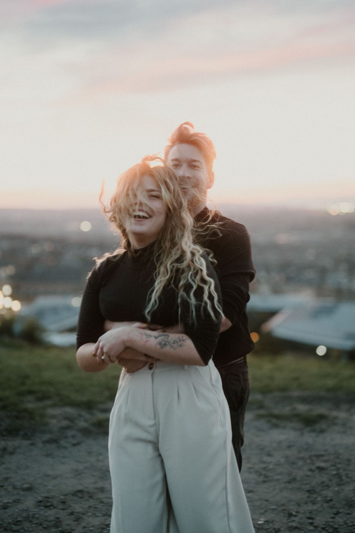 Couple embracing at sunset, laughing woman with curly hair in white pants, man hugging her from behind.
