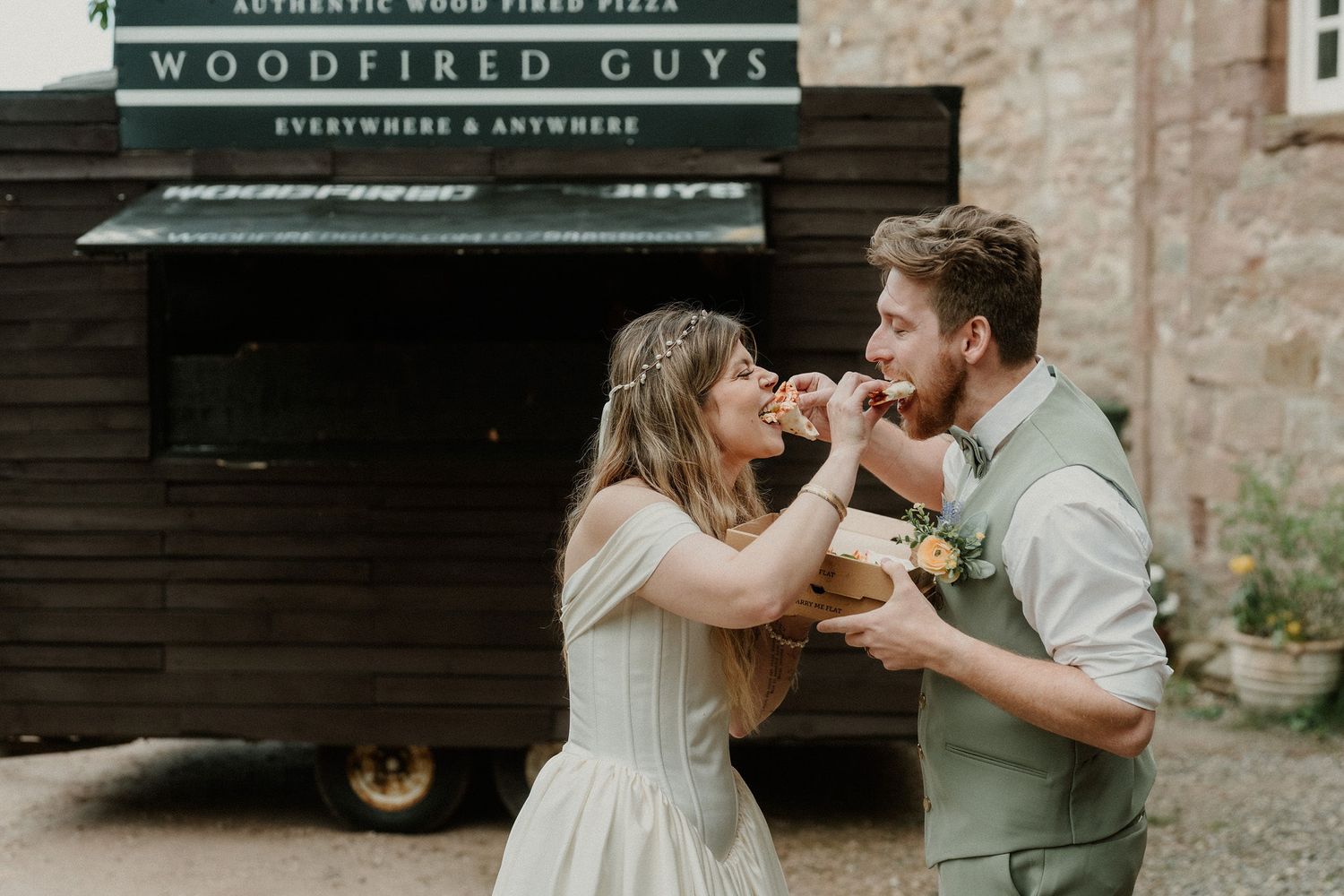 Bride and groom sharing pizza from Woodfired Guys food truck at rustic outdoor wedding celebration.