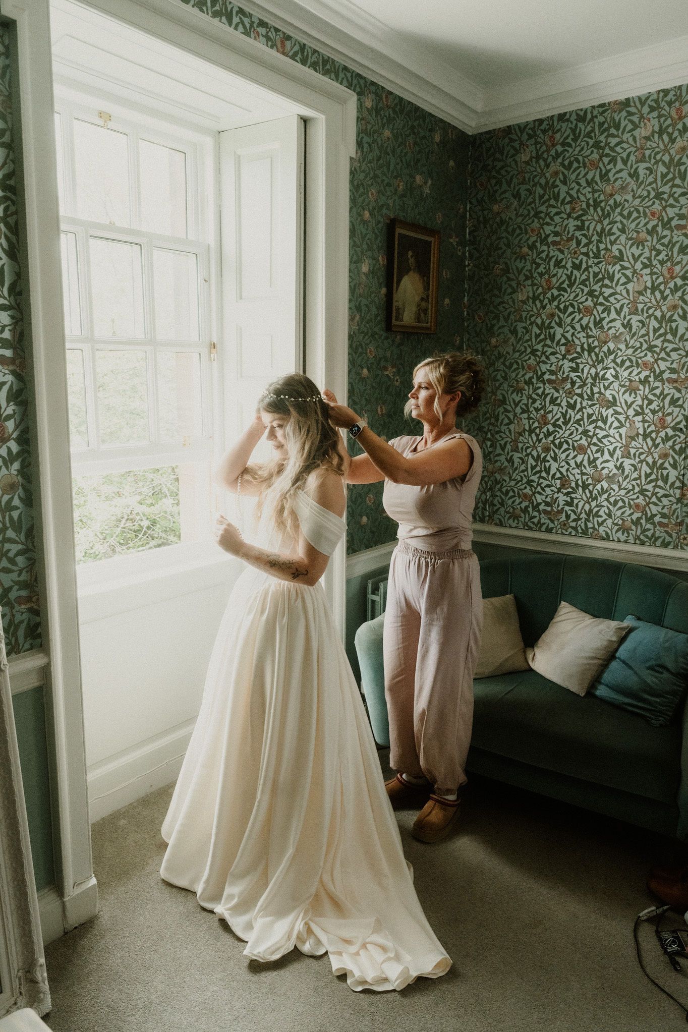 Bride in white wedding gown getting veil adjusted by bridesmaid near window in elegant green floral wallpapered room.