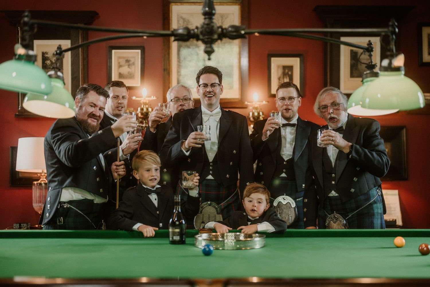 Groomsmen in formal black tuxedos and bow ties raise glasses around a green pool table with two young boys in matching suits.