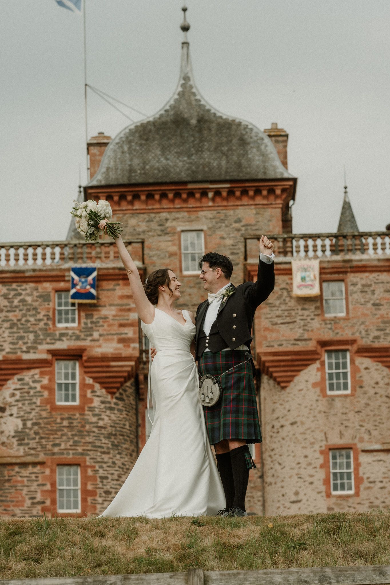 Bride in a white gown and groom in a kilt raise their arms in celebration in front of a historic red sandstone Scottish castle.
