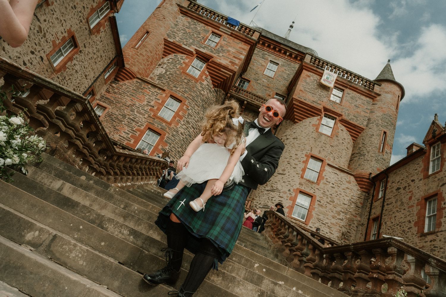 Groom in a green tartan kilt and bow tie carries a flower girl up stone steps at a historic red sandstone Scottish castle.