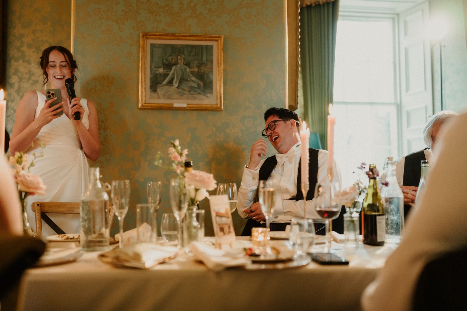 Woman in a white dress speaks into a microphone during a wedding reception toast as seated guests laugh at a elegantly set table.