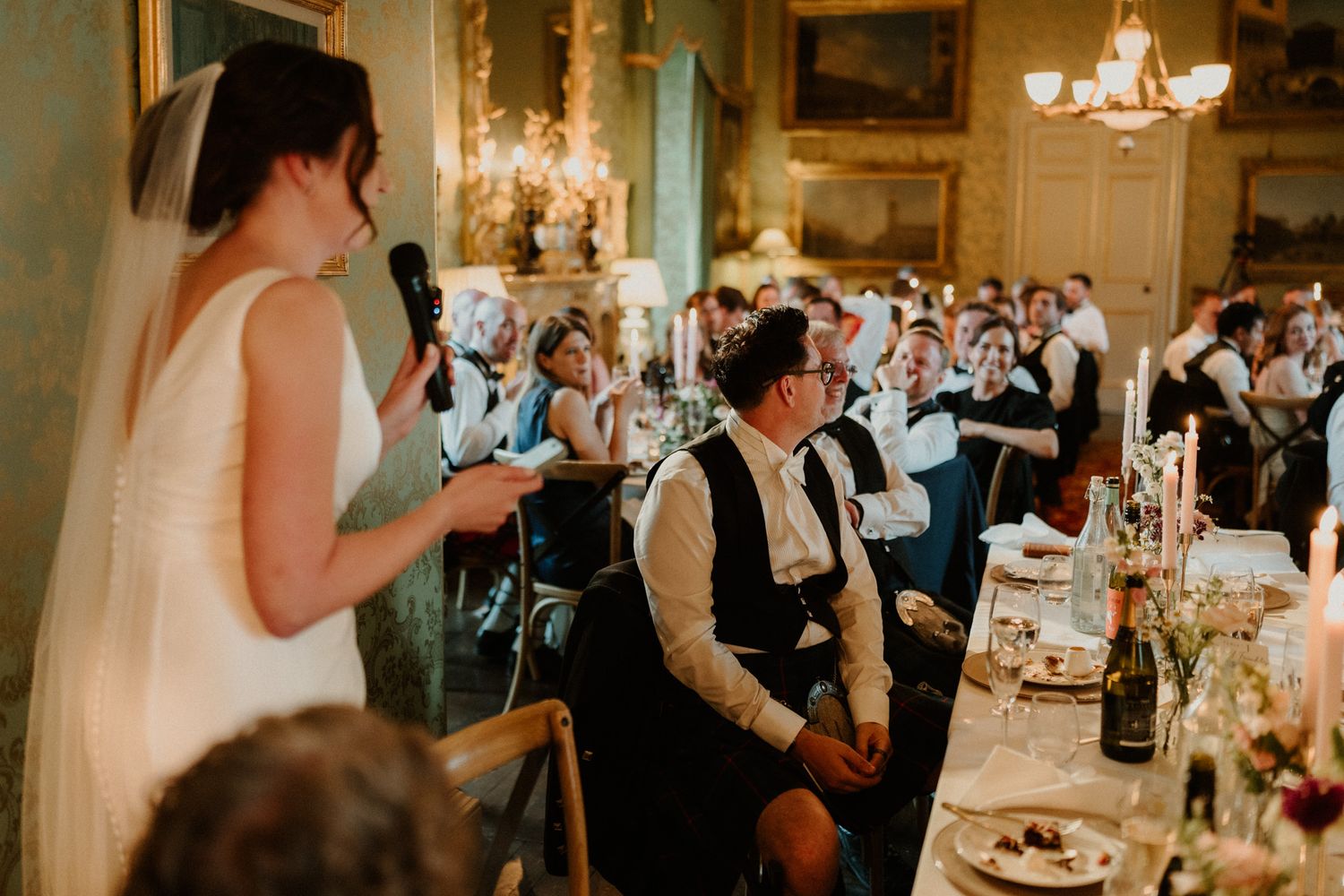 Bride in a white gown and veil gives a wedding speech with a microphone as seated guests listen at an elegant reception table.