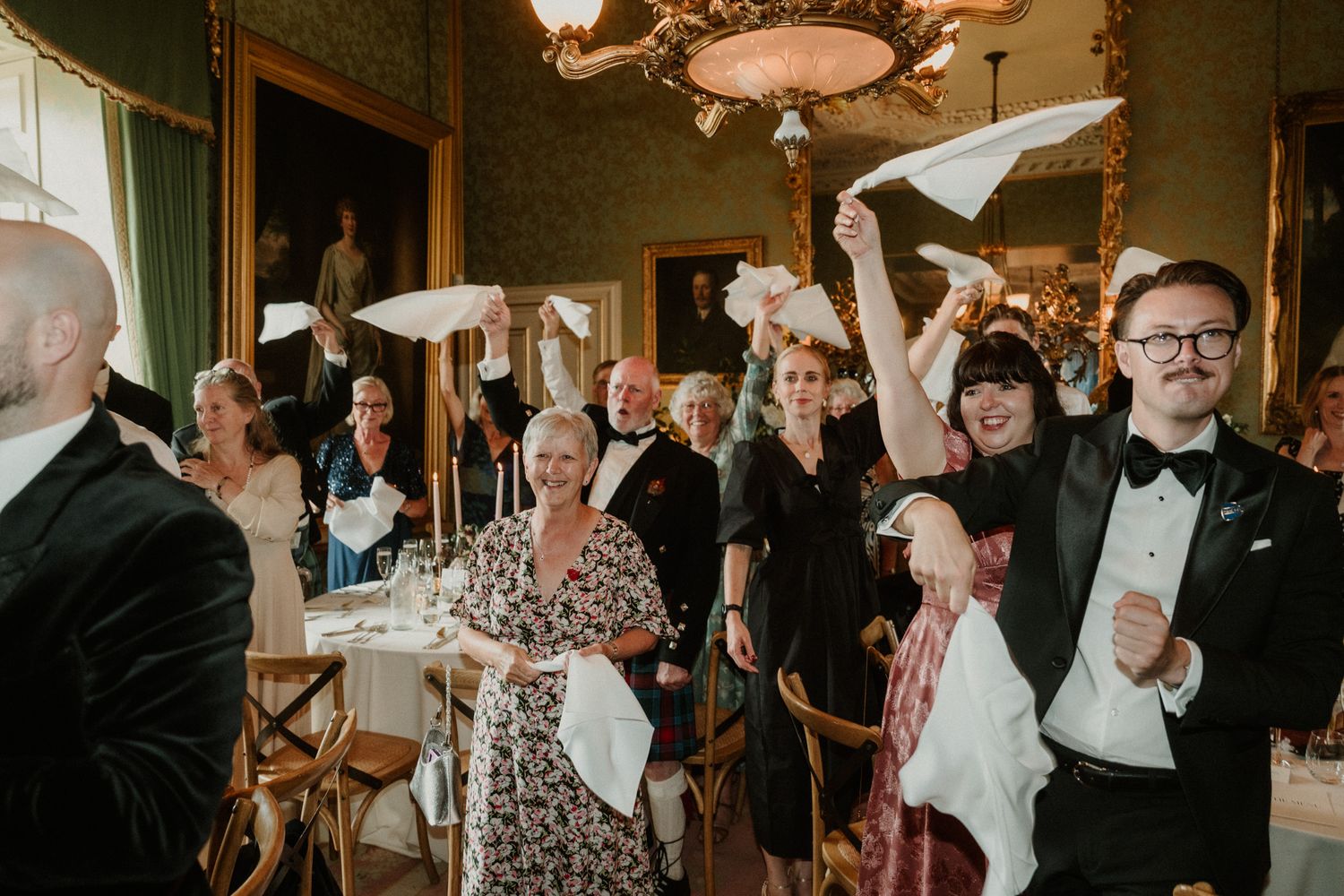 Wedding guests in formal attire wave white napkins and cheer in an elegant ballroom with a chandelier, as a man in a bow tie laughs.
