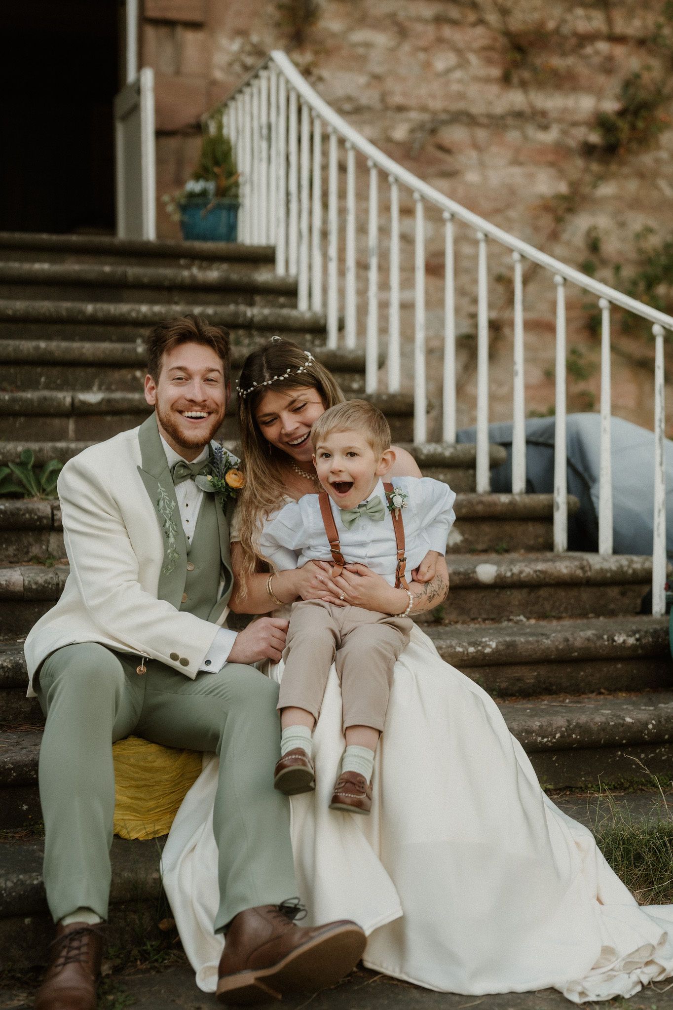 Happy bride and groom sitting on stone steps with their young child on wedding day, all smiling and laughing together.