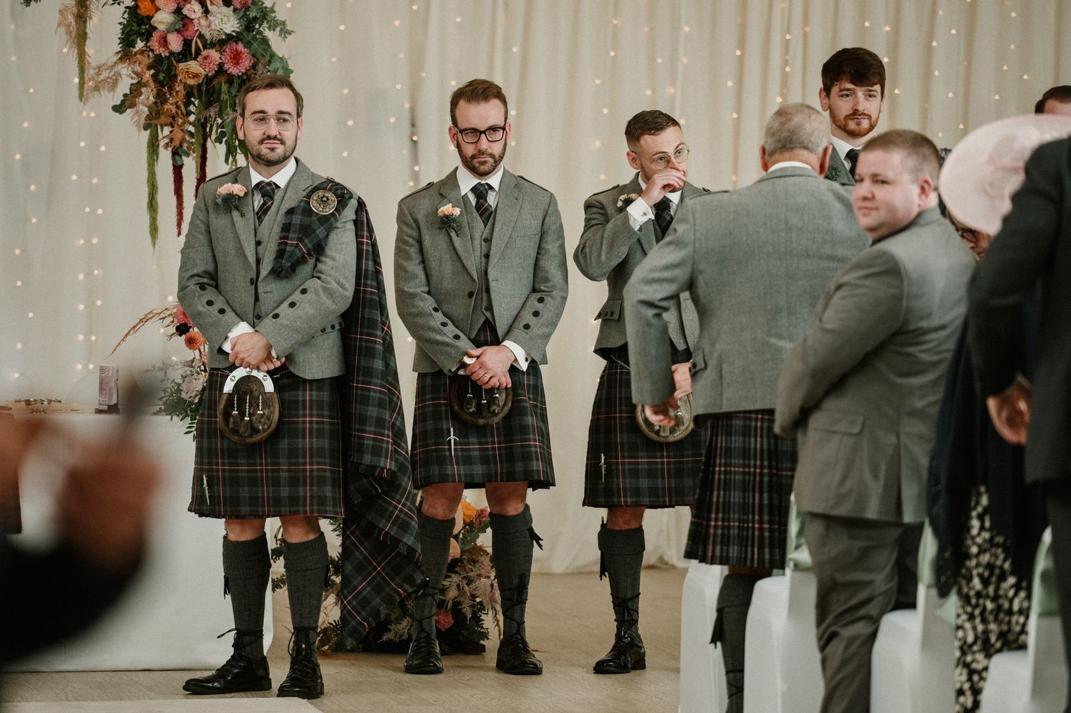 Groomsmen in traditional Scottish kilts and suits gather during wedding ceremony.