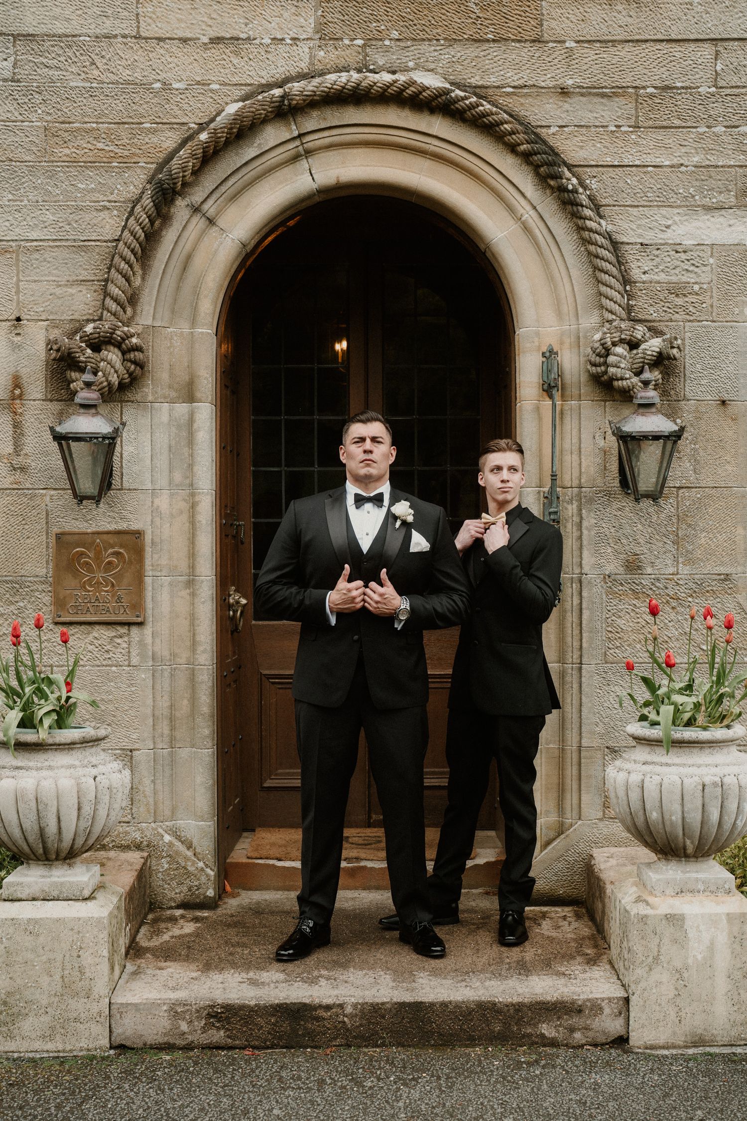 Two people in tuxedos standing in front of an ornate stone archway with decorative planters.