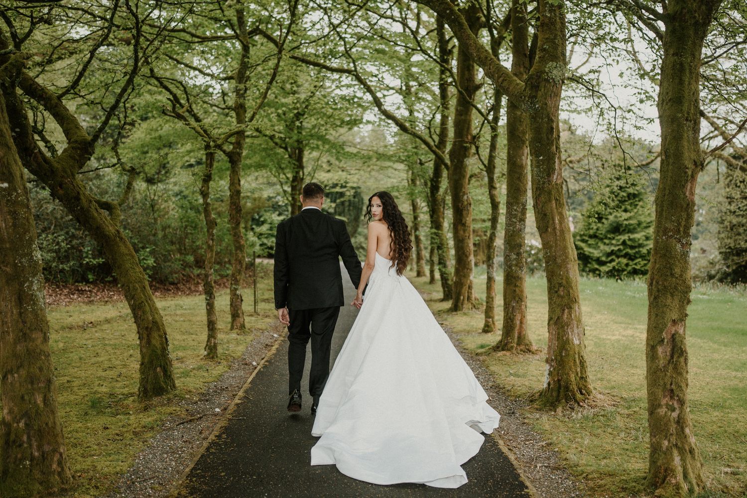 A wedding couple walks through a tree-lined path creating a romantic woodland scene.