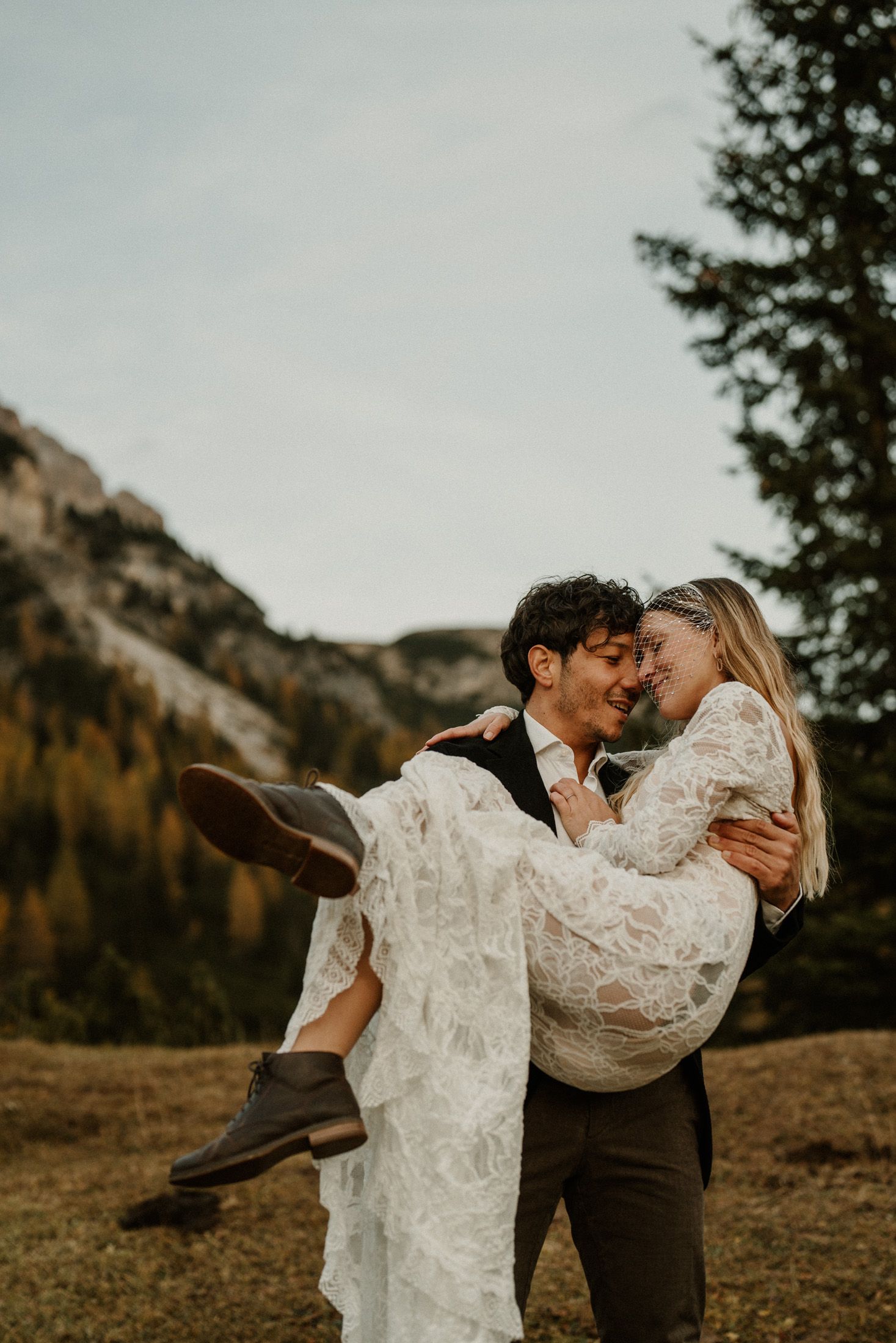A couple shares a romantic embrace against a backdrop of towering mountain peaks and autumn trees.