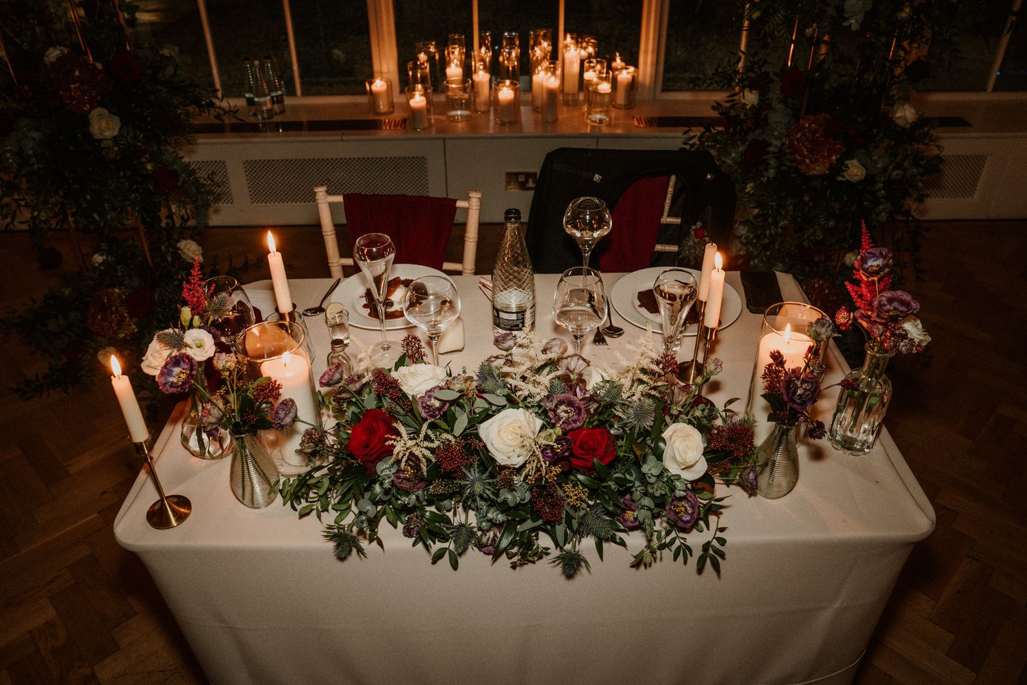 Elegant wedding reception table decorated with candles, red flowers and greenery.