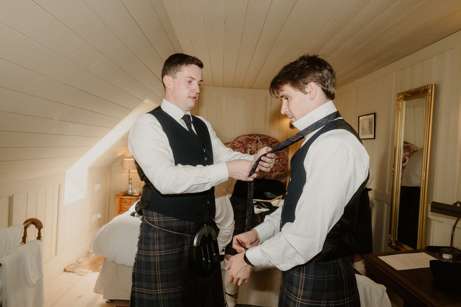 Two groomsmen in kilts and black vests help each other with neckties while getting ready for a Scottish wedding.