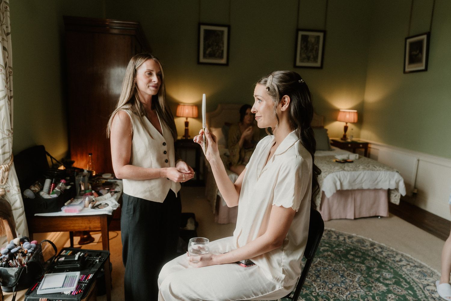 A makeup artist applies finishing touches to a bride seated in an elegant green-walled bedroom before her wedding.