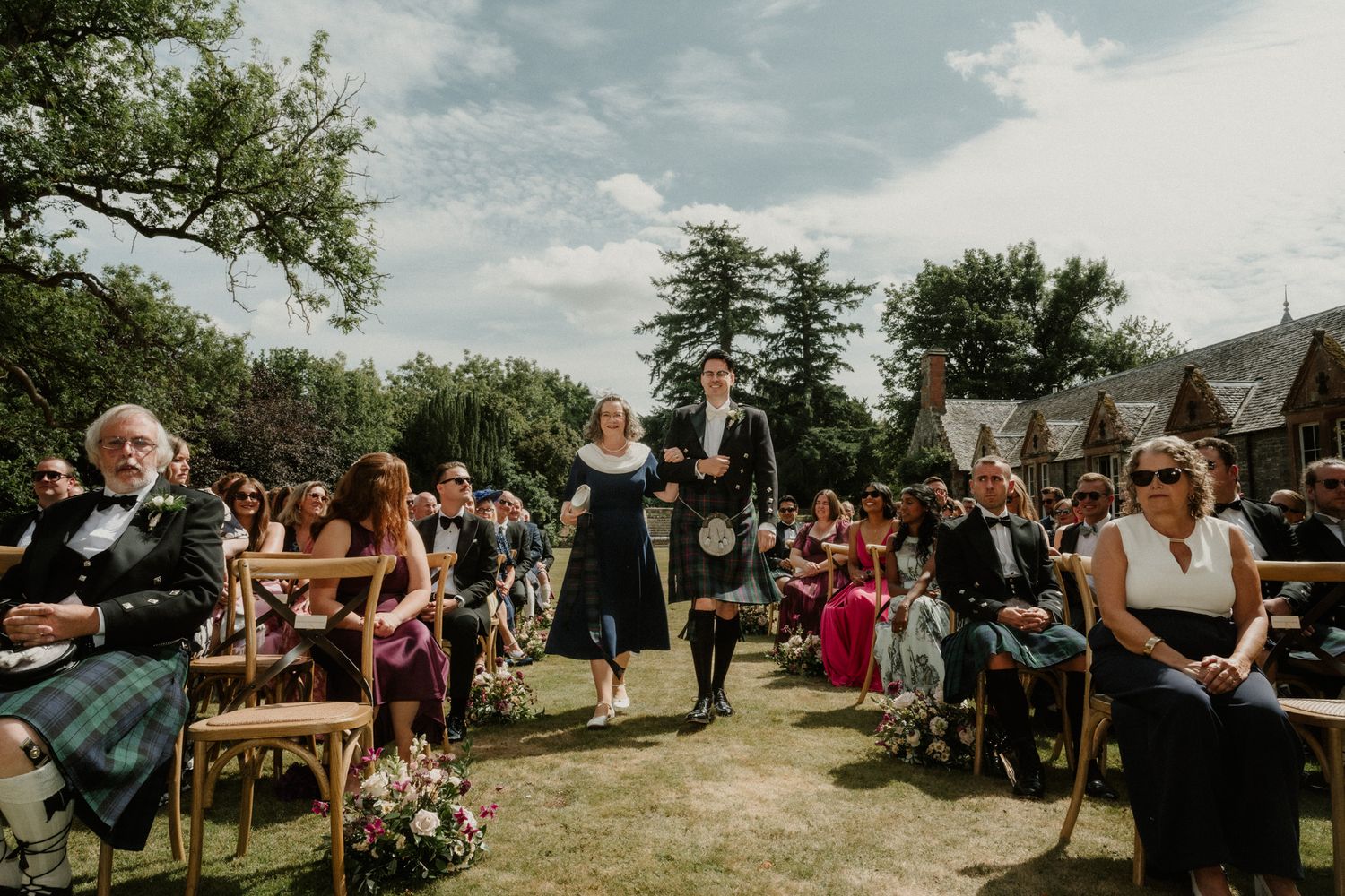 Wedding guests in kilts and formal attire sit on wooden chairs as a couple walks down the grass aisle at an outdoor ceremony.