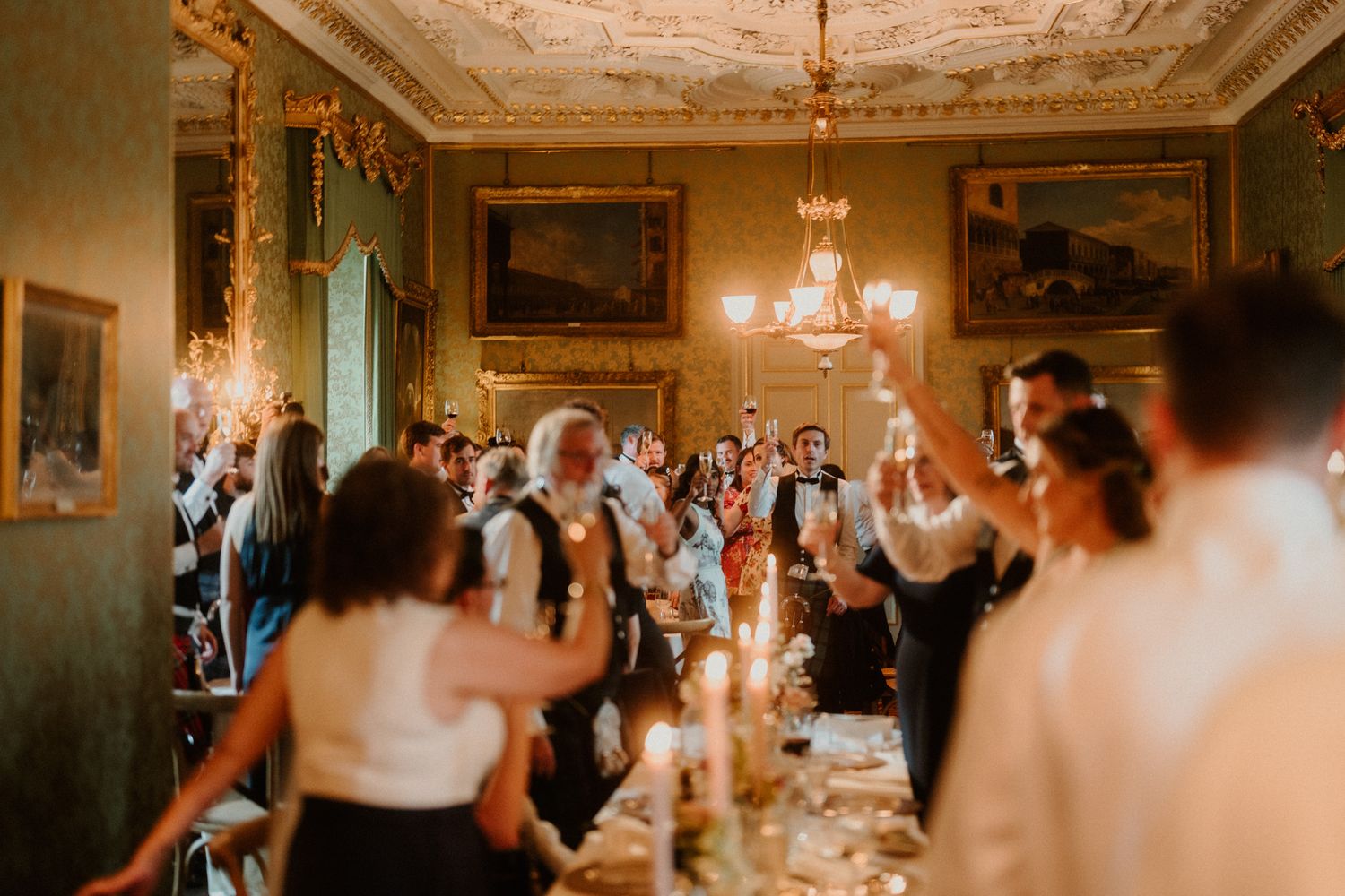 Wedding guests raise their arms and cheer around a candlelit table in an elegant historic room with ornate ceilings and a chandelier.