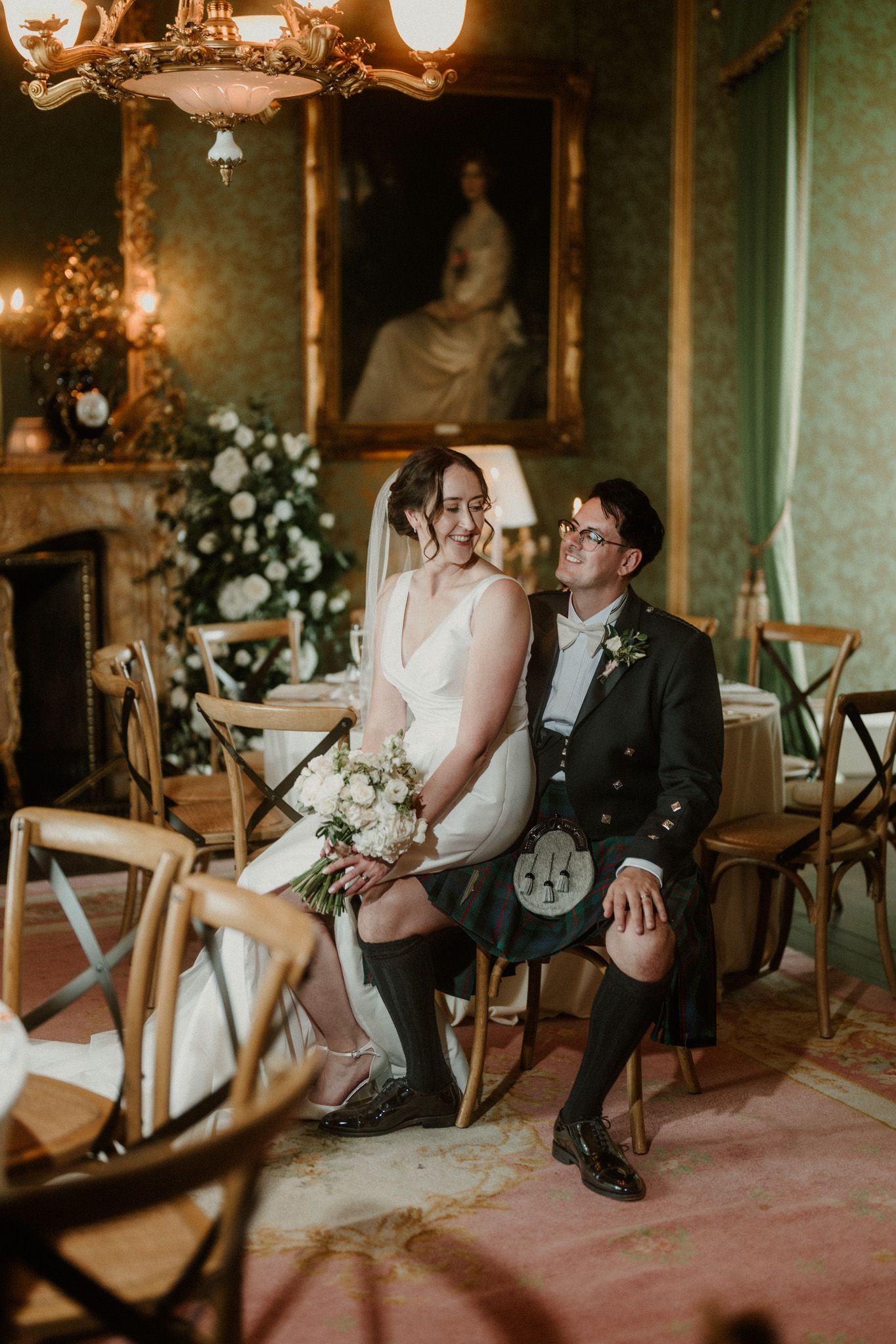 Bride in a white wedding dress and groom in a Scottish kilt and jacket sit smiling together in an elegant room with floral arrangements.