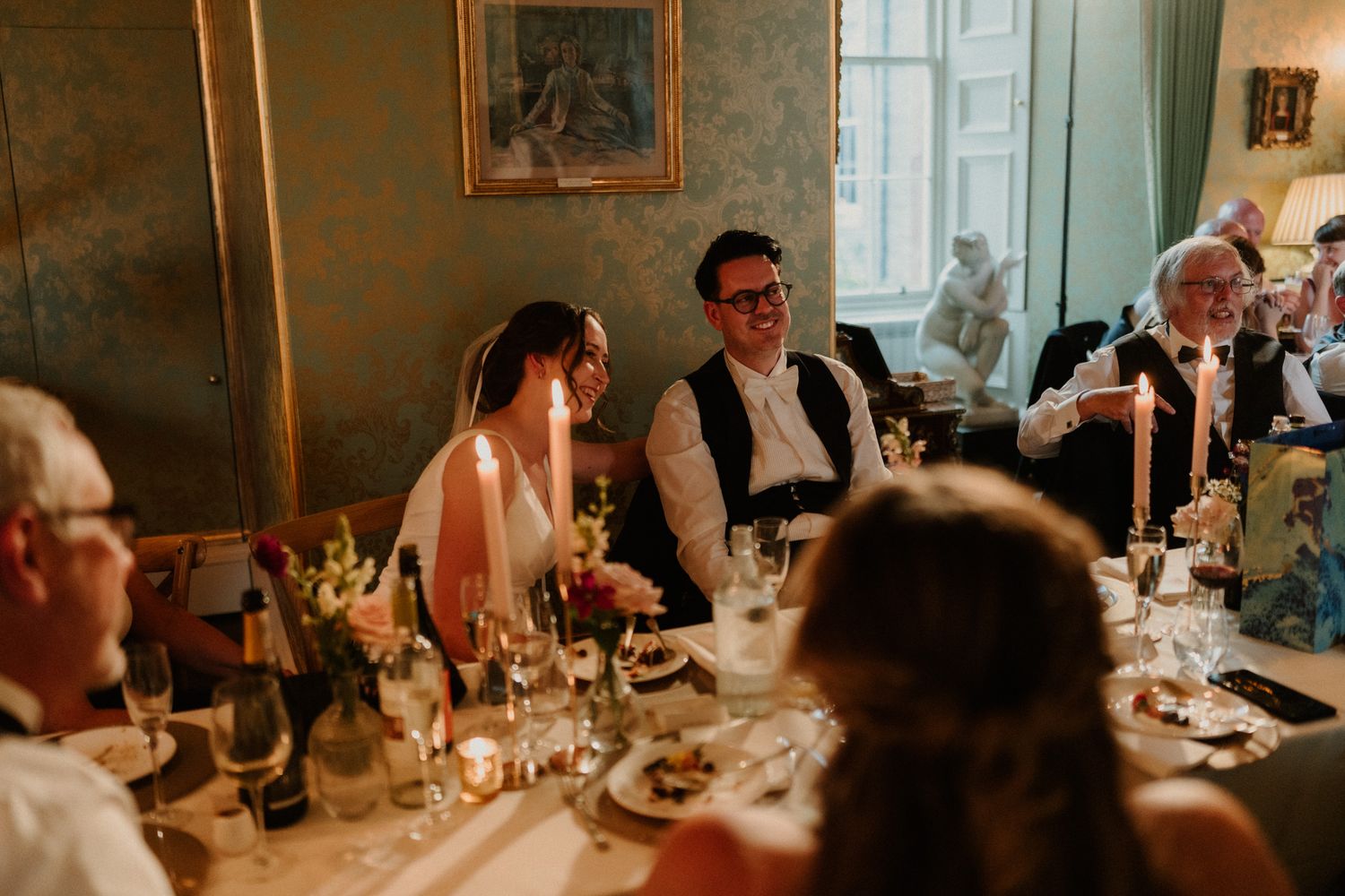 Smiling wedding guests laugh together at a candlelit dinner table with flowers, wine glasses and plates in an elegant room.