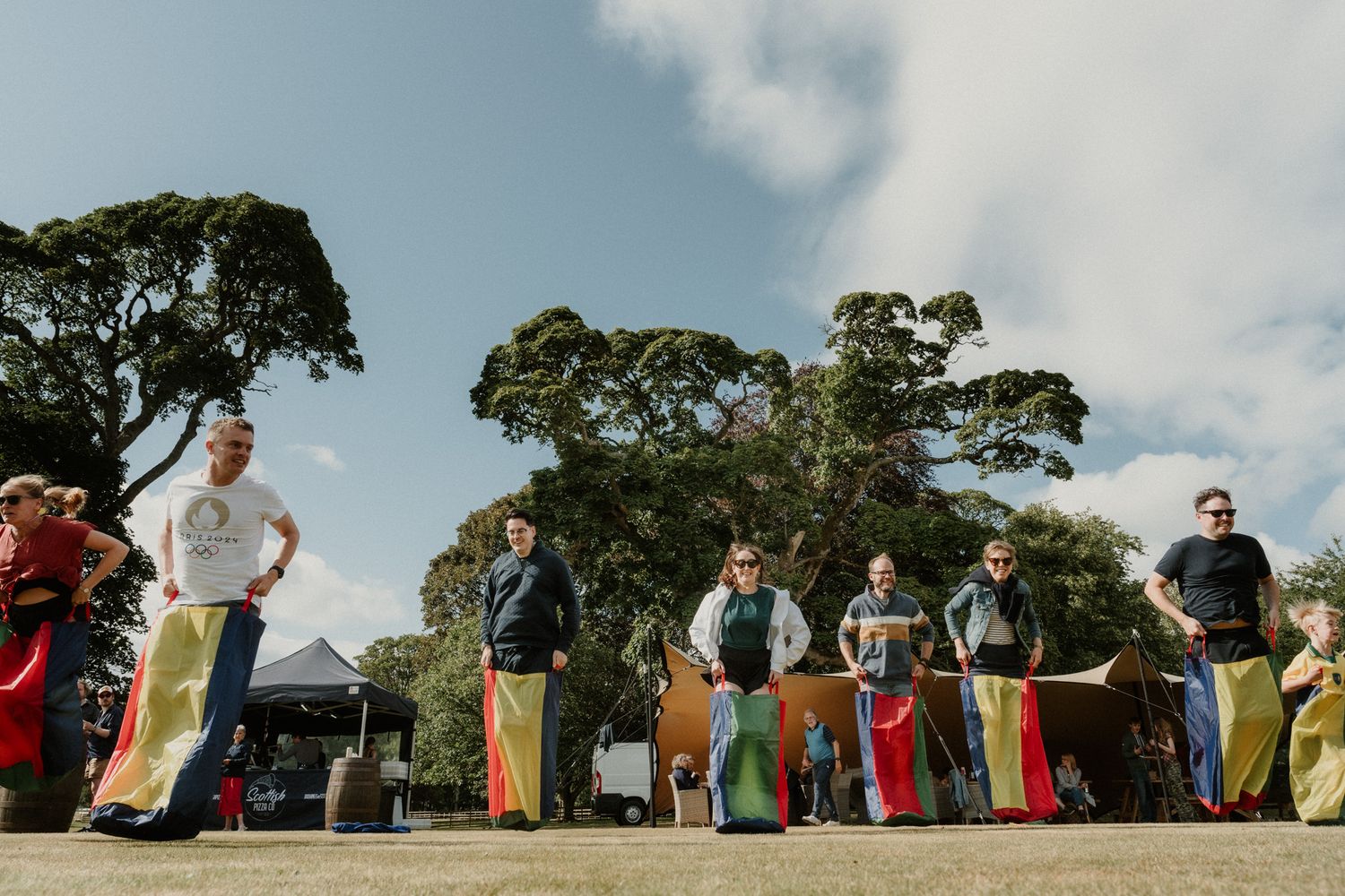 People line up outdoors holding colorful sacks at a sack race event in a sunny park with large trees and a blue sky.