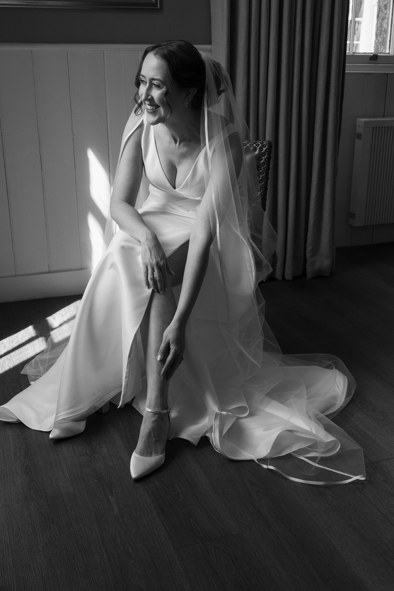 Smiling bride in an elegant gown and long veil sits by a sunlit window, adjusting her pointed bridal shoe in a black and white photo.
