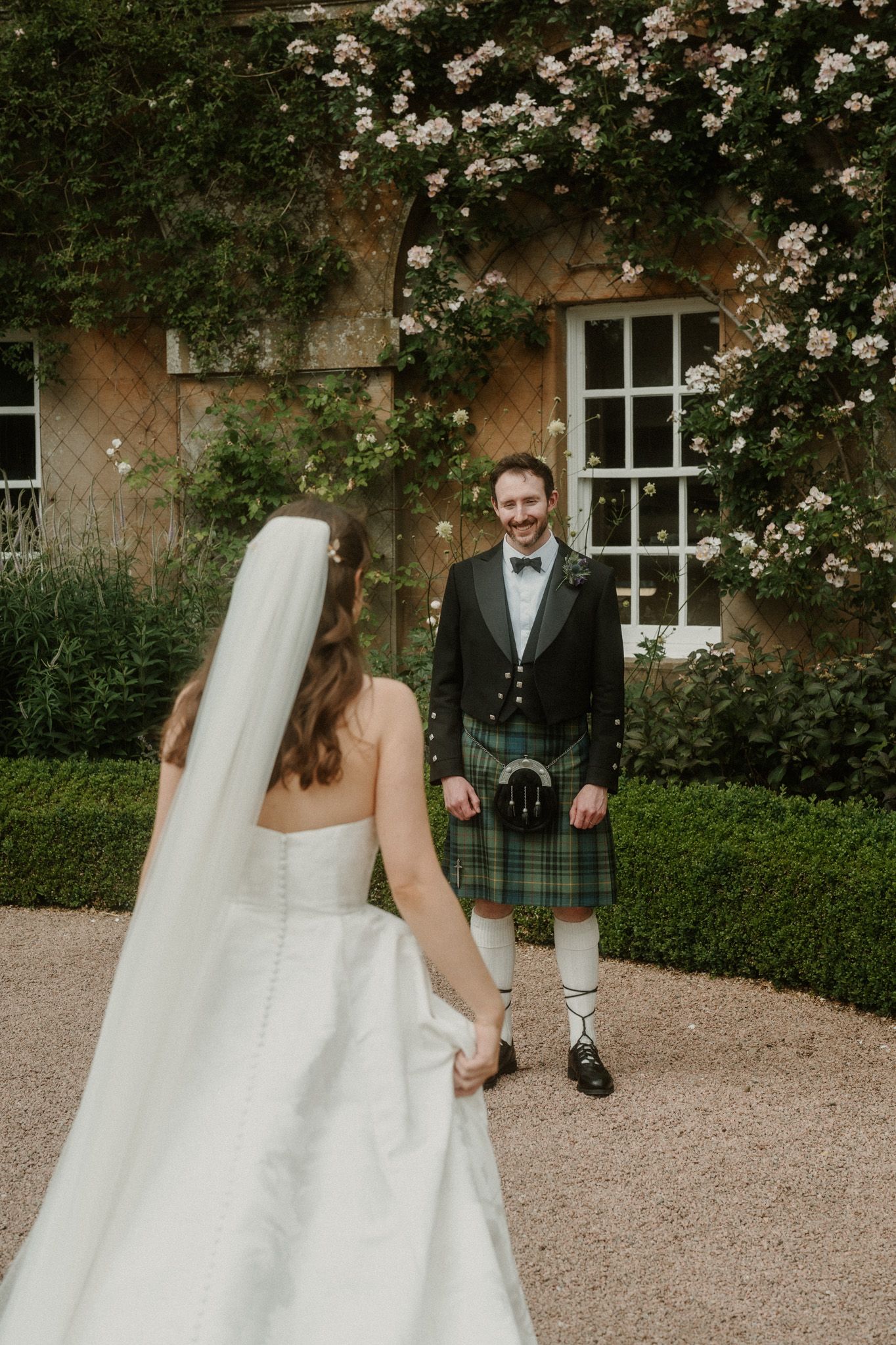 Bride in white gown and veil walks toward smiling groom in Scottish kilt during outdoor wedding ceremony.