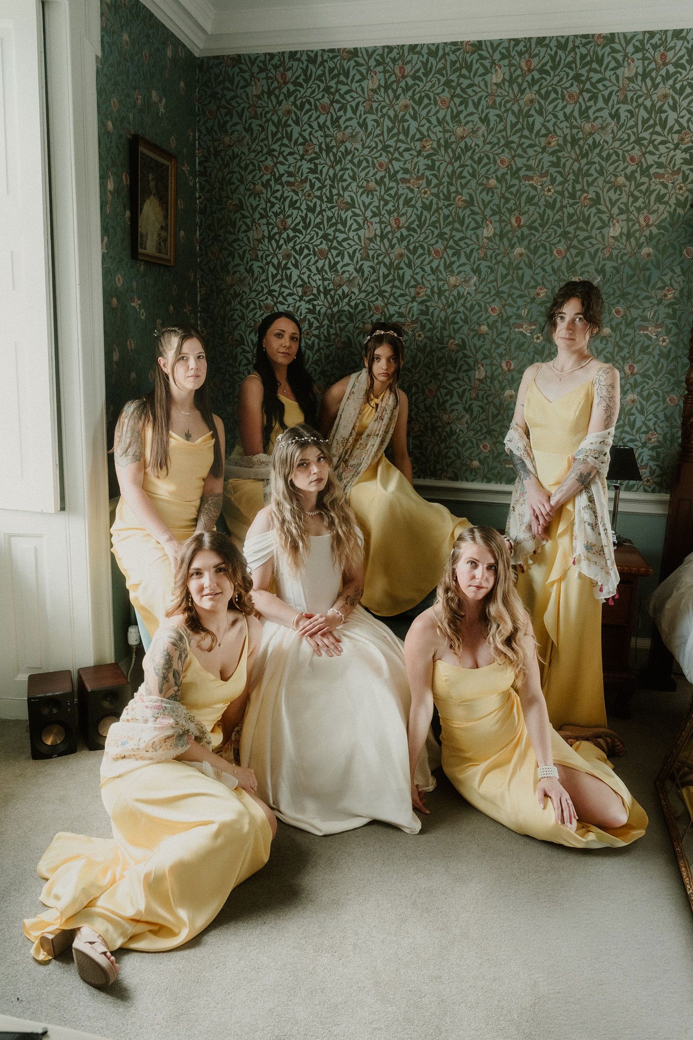 Bride in white gown poses with six bridesmaids in yellow satin dresses in a room with green floral wallpaper.
