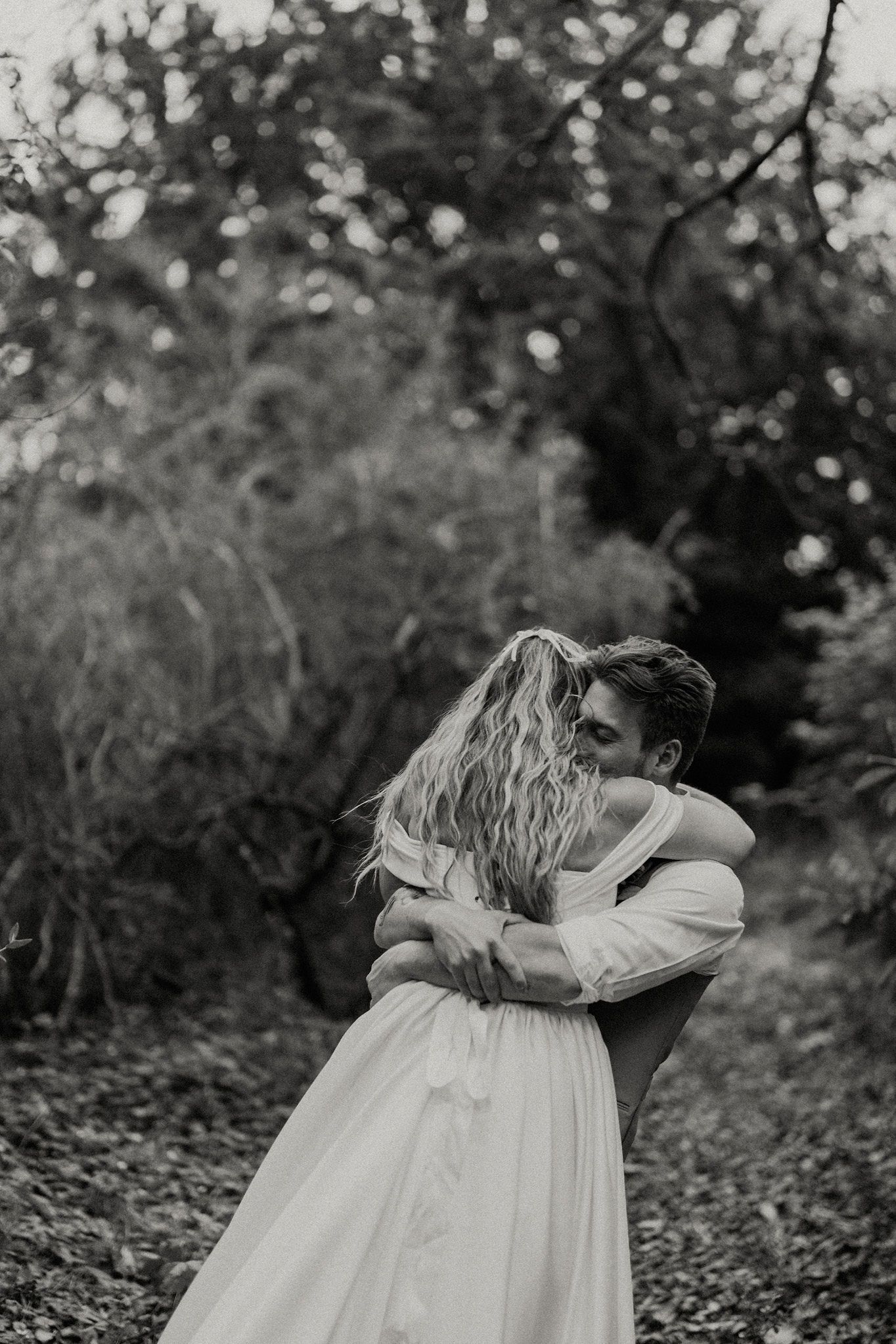 Black and white photo of a couple embracing in a forest, bride in flowing white dress hugging groom outdoors.