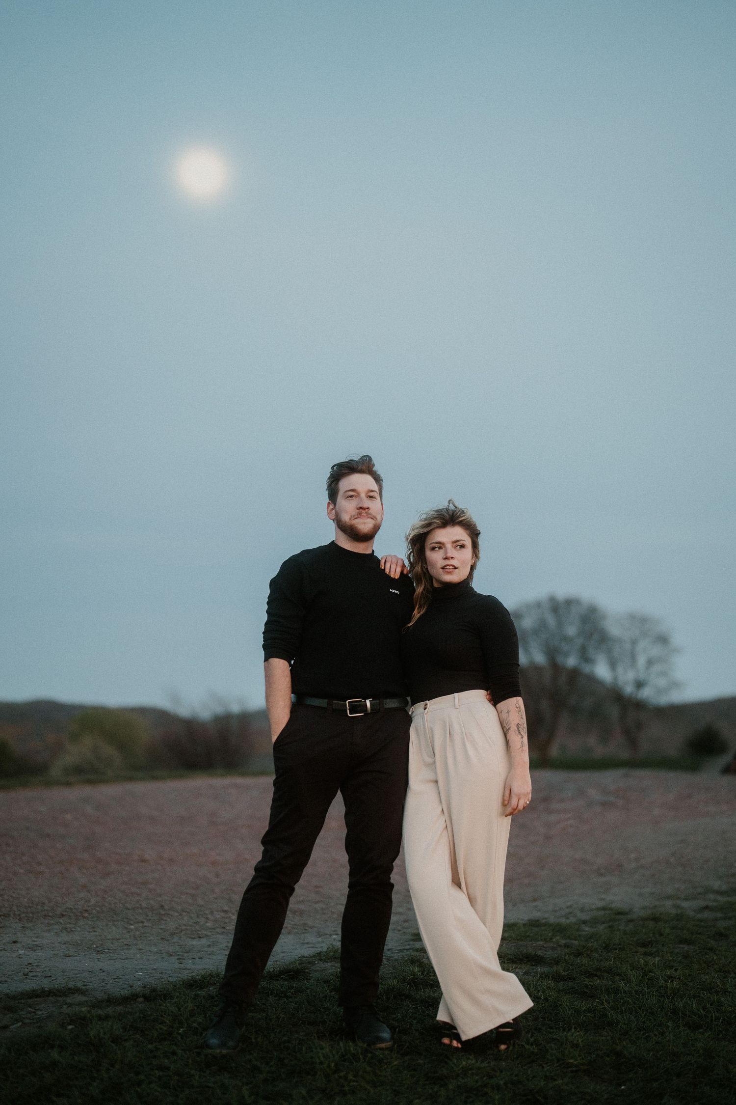 Couple posing together outdoors at dusk with a full moon glowing in the blue sky behind them.