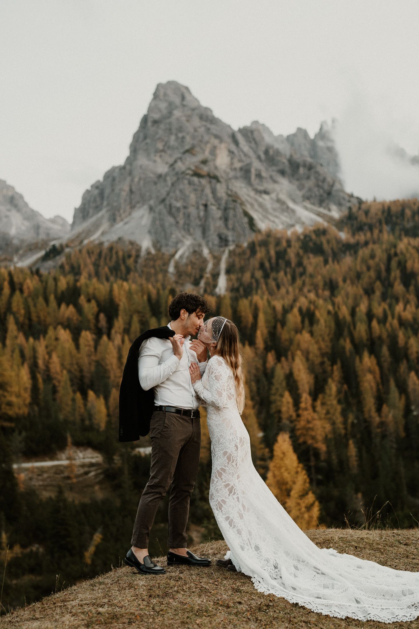 Couple sharing a kiss in wedding attire with dramatic Dolomites mountains and autumn forest backdrop.