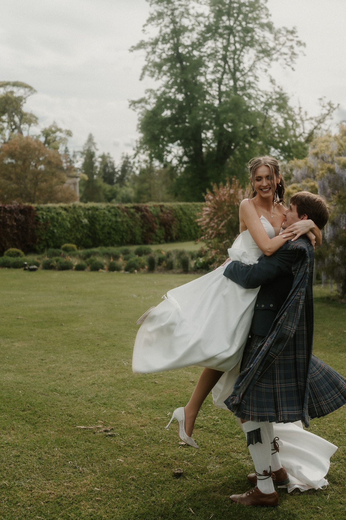 Groom in tartan kilt lifts smiling bride in white wedding dress outdoors in lush green garden setting.