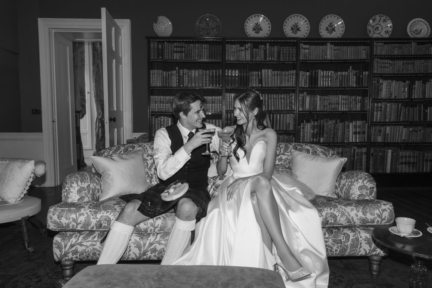 Black and white photo of a bride and groom toasting in a cozy library, seated on a floral sofa surrounded by bookshelves.