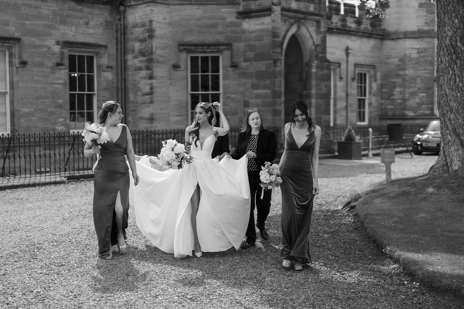 Bride in flowing white gown walks with bridesmaids in dark dresses outside a stone building in black and white photo.