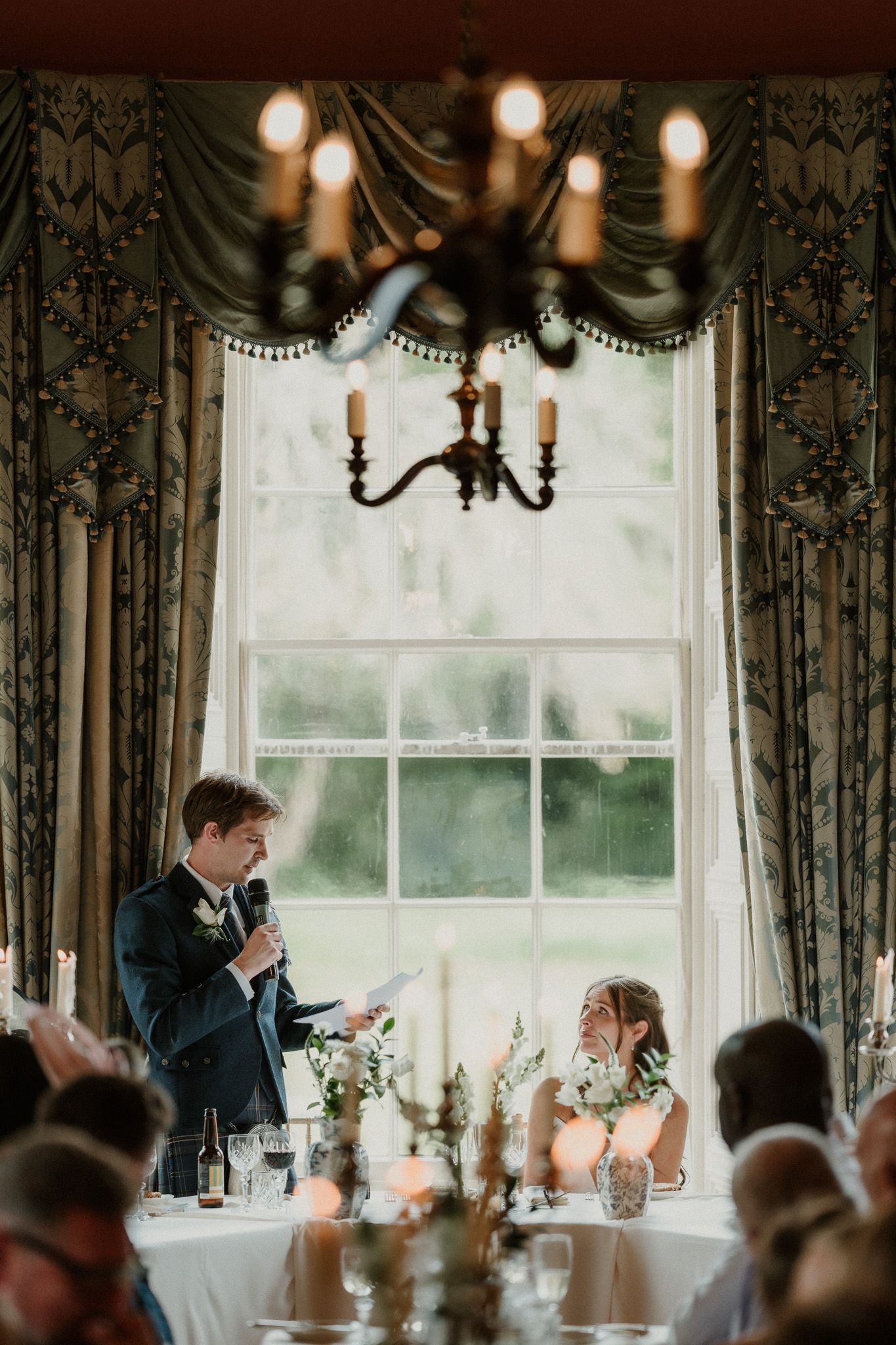 Groom delivers wedding speech at candlelit reception table beneath chandelier, bride smiling beside floral centerpieces.